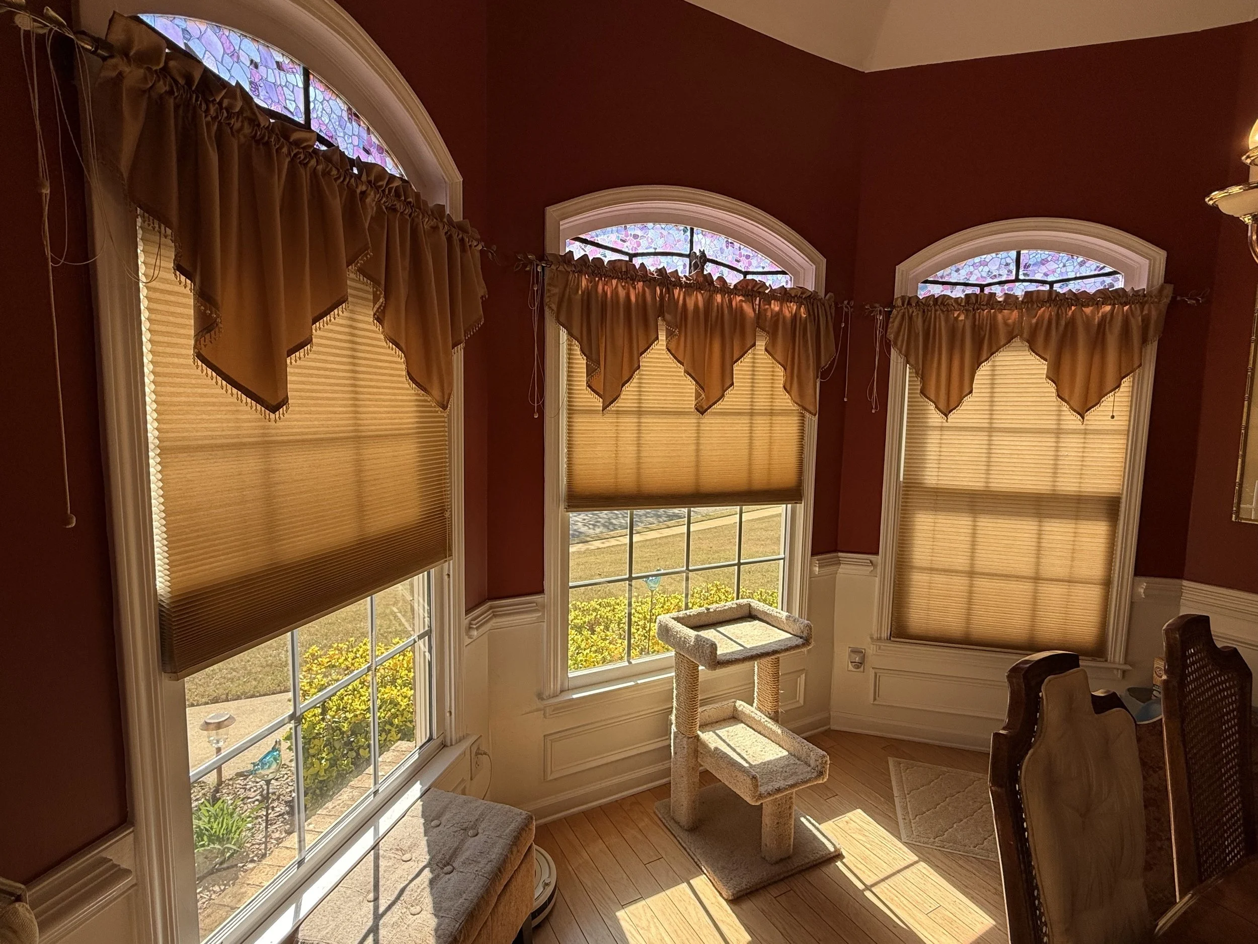 Sunlit room with three large arched windows covered with beige blinds and matching valances. A cat tree stands in front of the central window, and part of a chair and a bench are visible in the room.