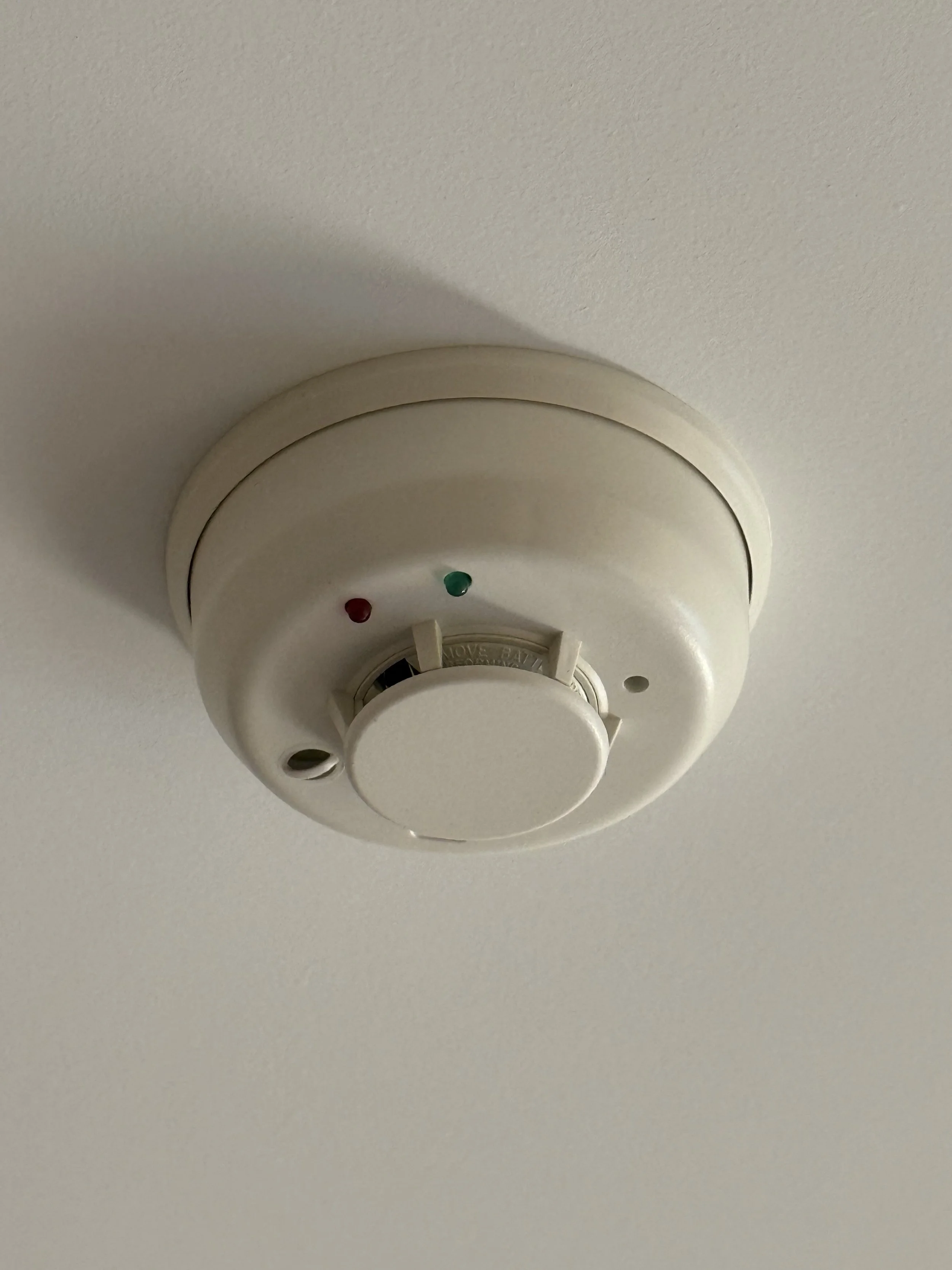 A white ceiling smoke detector with red and green indicator lights mounted on a white ceiling.