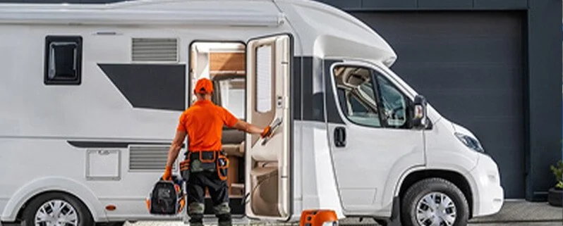 A person in orange with a tool bag enters a white camper van parked outside a building.