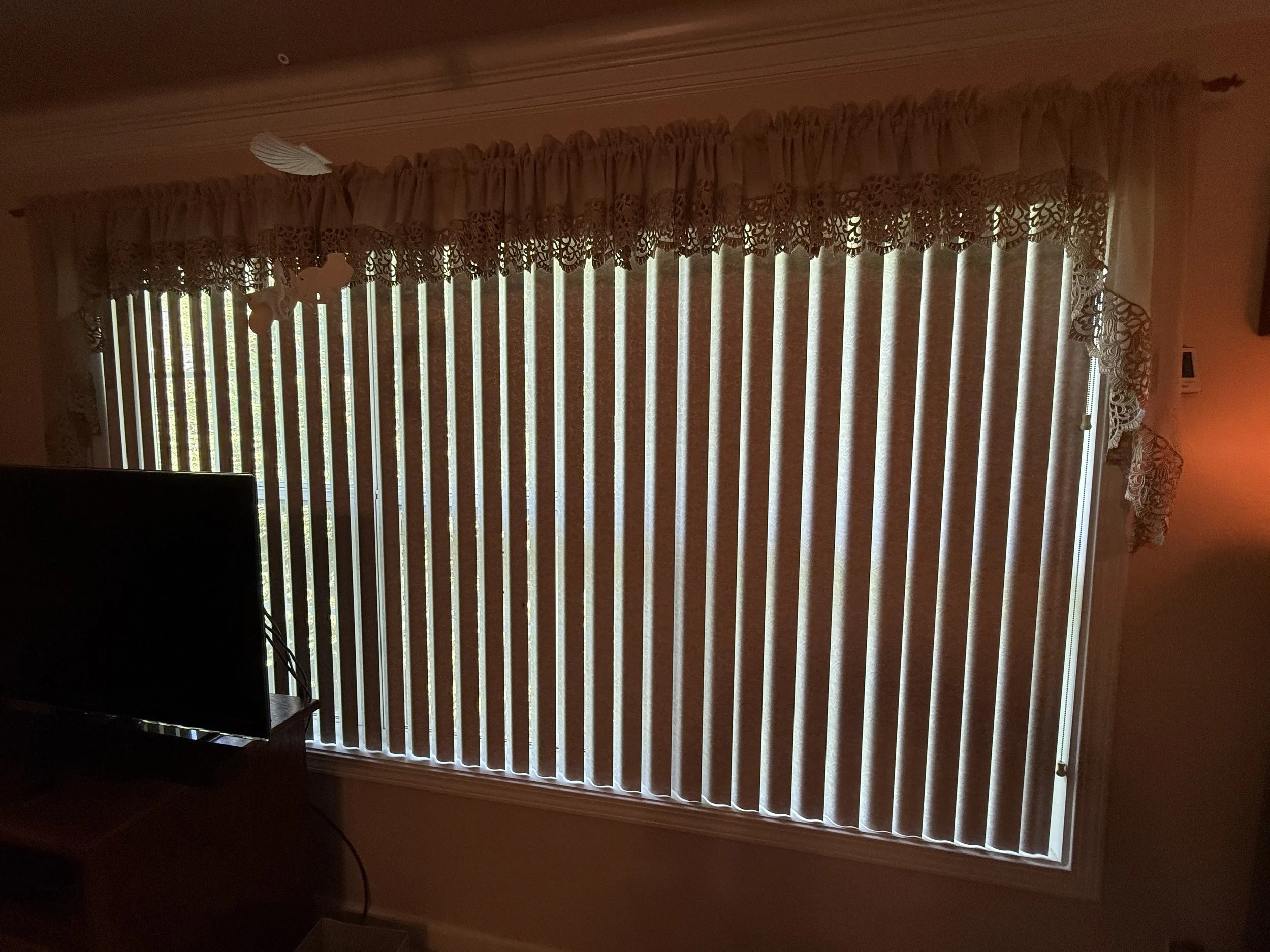 A window with vertical blinds and lace curtains, with some light coming through. There is a TV in the foreground.