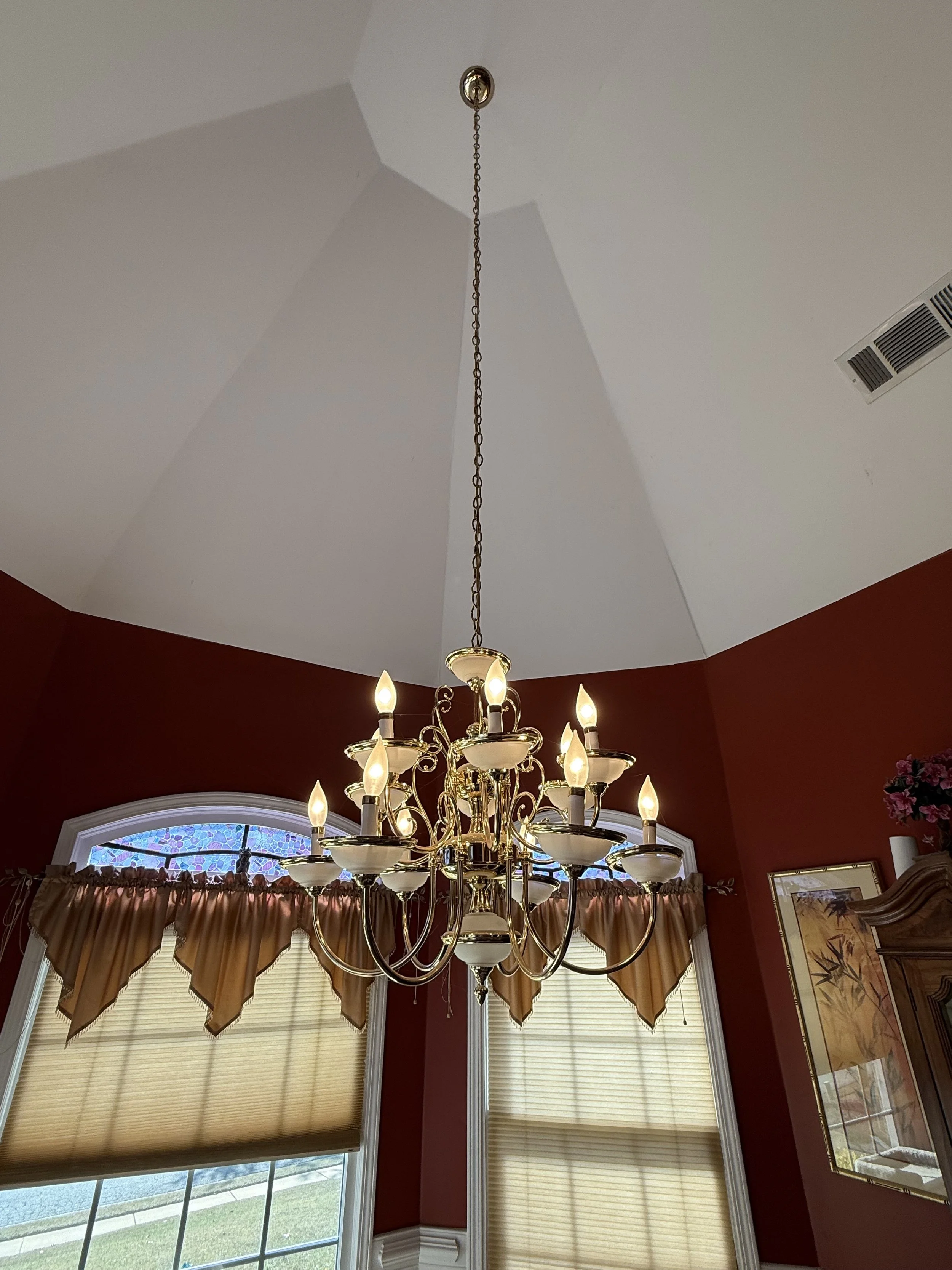 A chandelier with multiple candle-shaped light bulbs is hanging from a ceiling in a room with red walls, arched windows with beige blinds, and framed artwork on the wall.