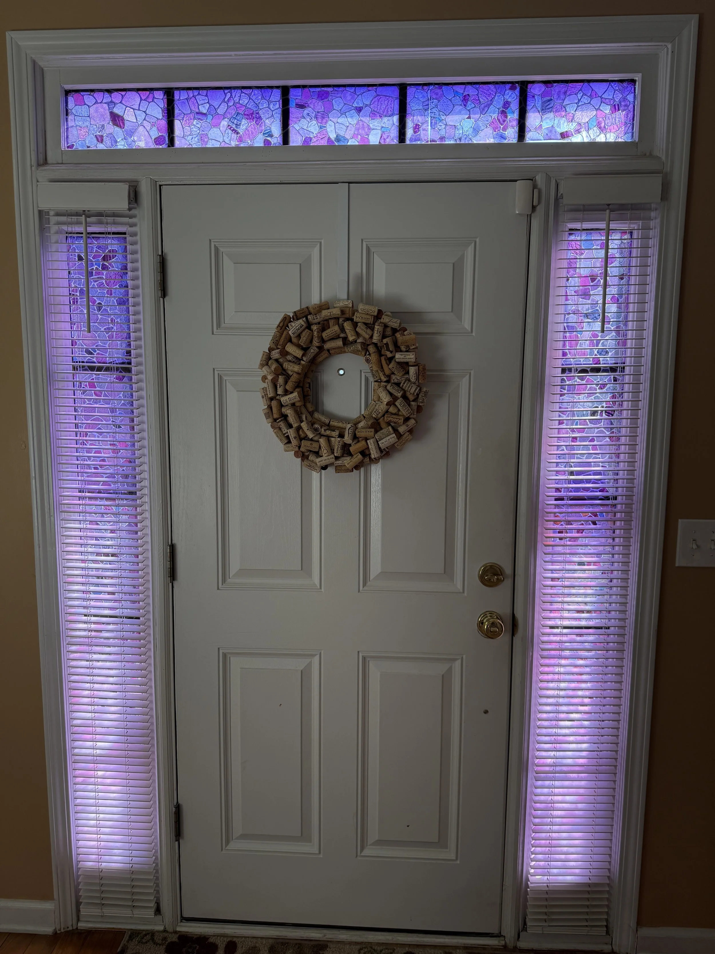 A white front door with a wreath made of wine corks hanging on it. The door has two panels and is flanked by vertical stained glass panels with purple and pink mosaic patterns, along with a horizontal stained glass transom window above, all illuminated with purple lighting.