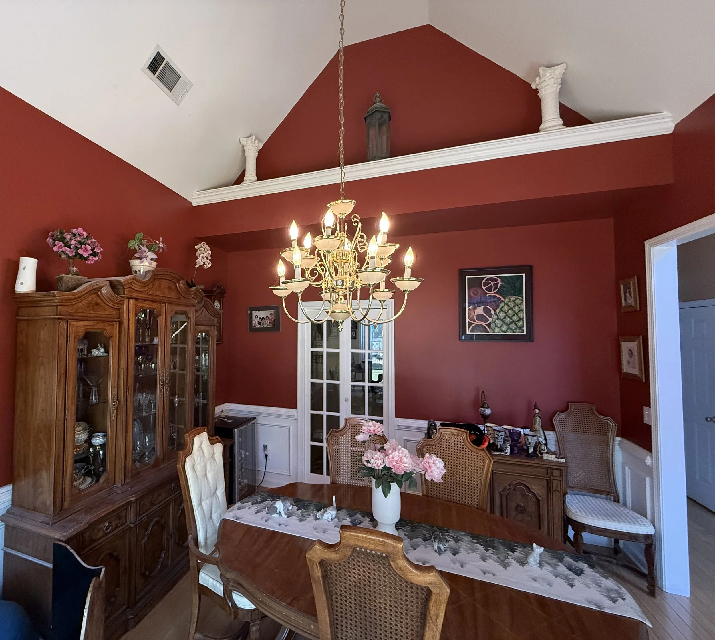 A dining room with red walls, a wooden table, and chairs. A large white vase with pink flowers is on the table. An ornate chandelier hangs from the ceiling, and a wooden cabinet is against the wall with decorative items and flowers. Framed artwork and a glass door are also visible.