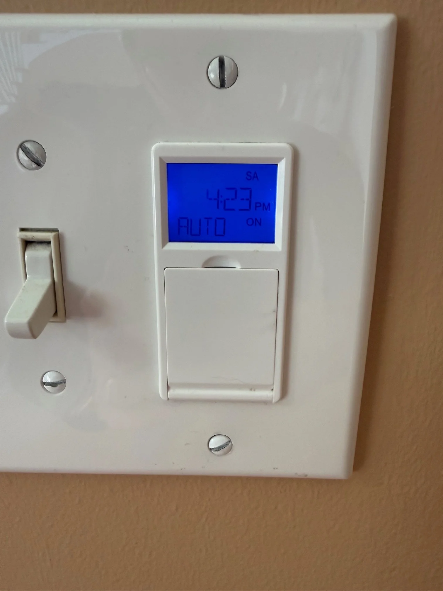 Close-up of a white light switchplate with a digital clock displaying 4:23 PM, on a beige wall.