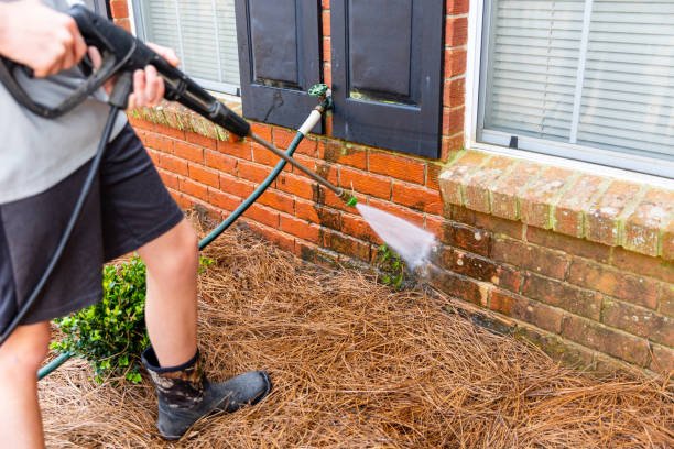 Person using a pressure washer to clean the exterior brick wall of a house, near a window with black shutters and a brick ledge.
