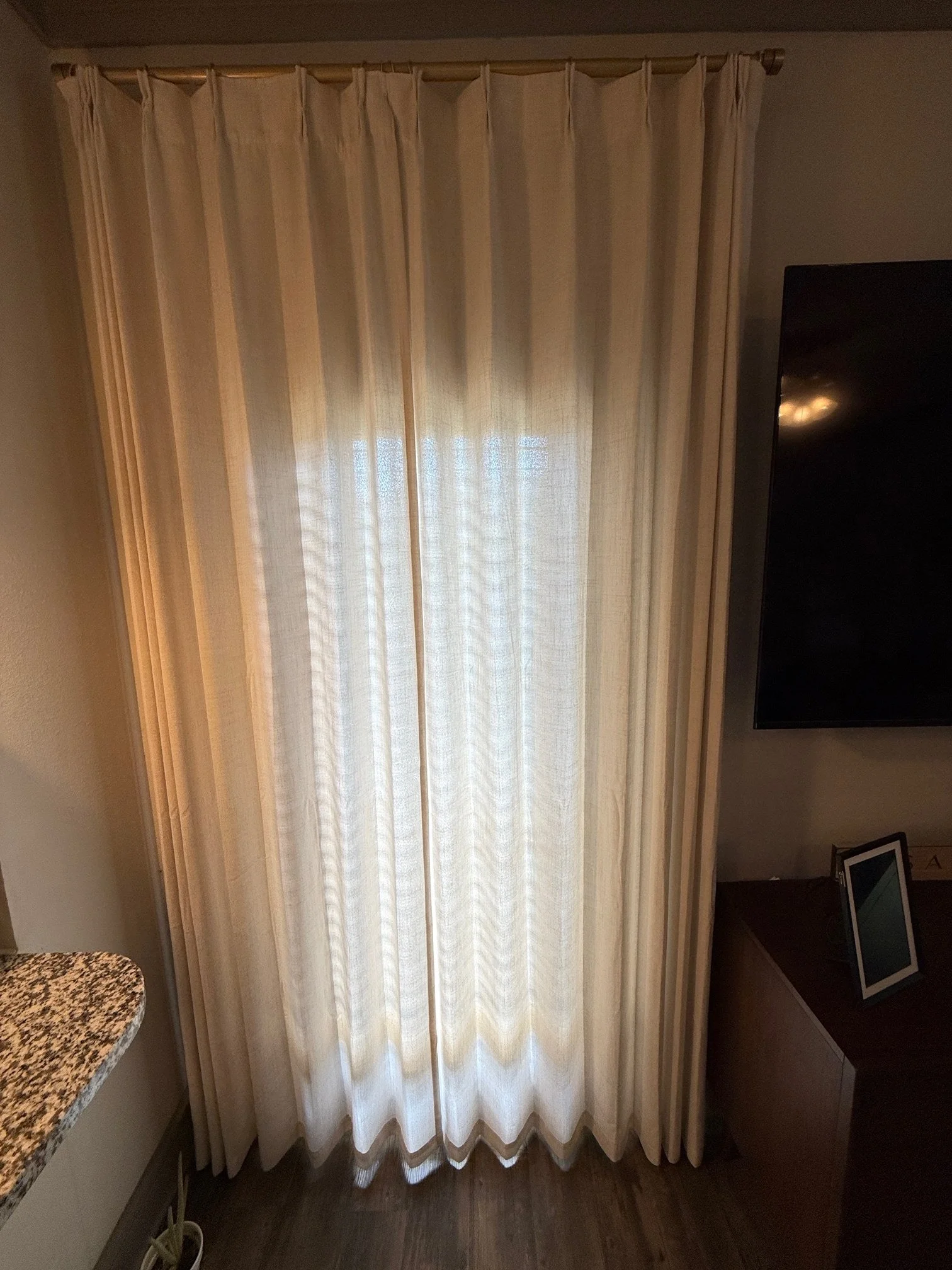 Cream-colored curtains hanging from a metal rod in front of a window, with a dark floor and part of a granite countertop visible on the left.