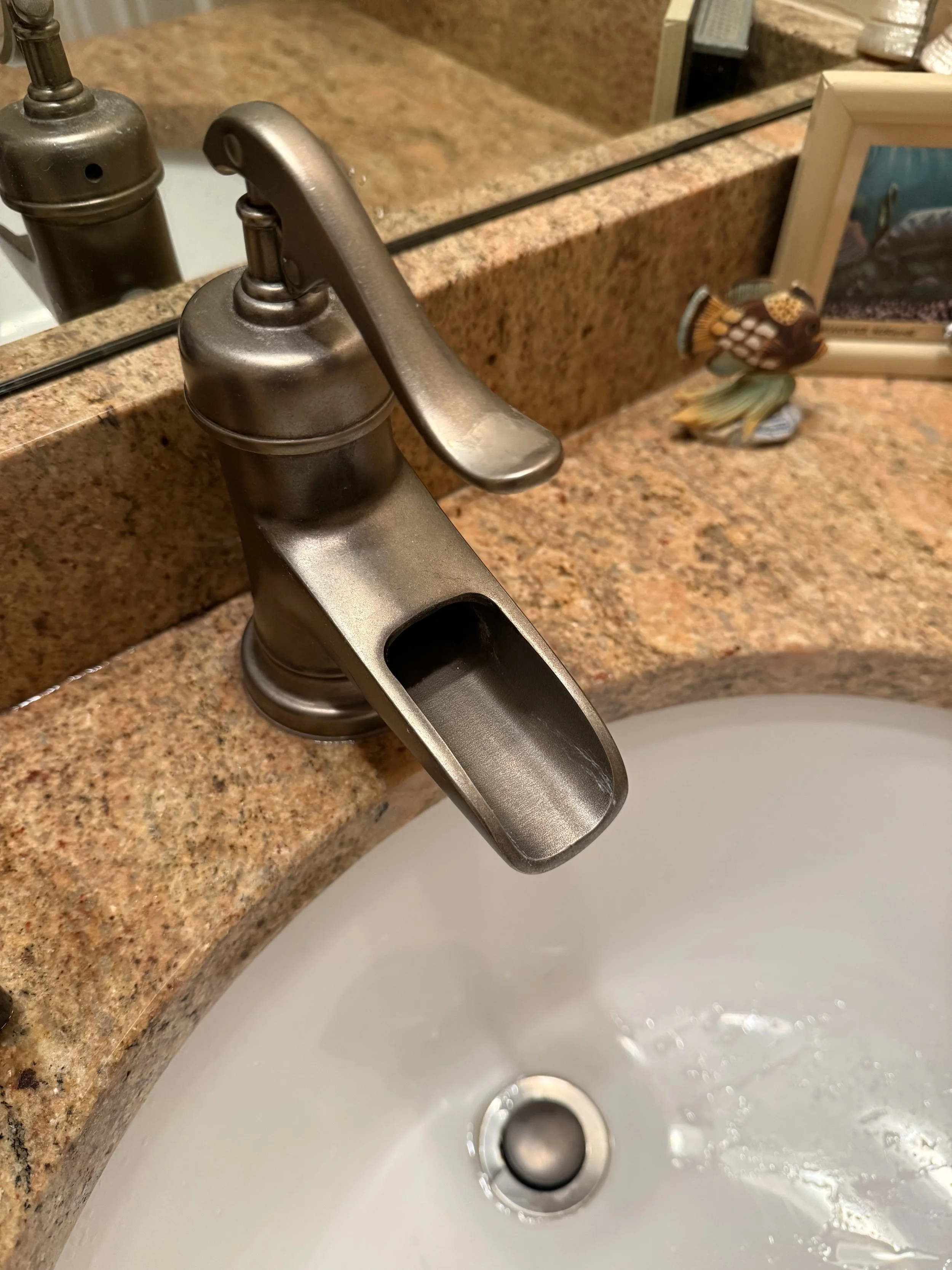 Close-up of a kitchen faucet over a sink with water running, granite countertop in the background, and a framed photo and decorative fish ornament on the counter.