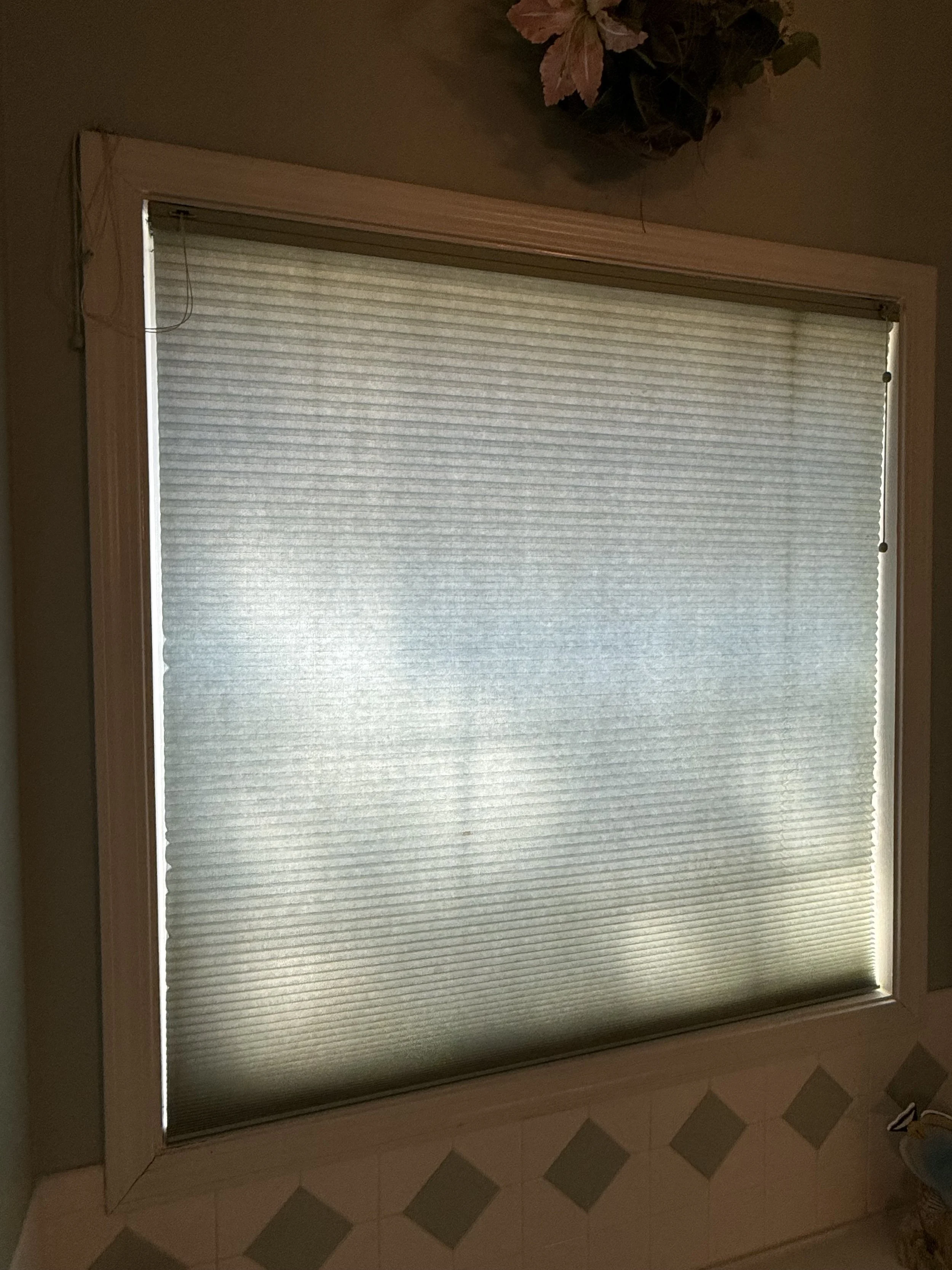 A window with a pleated screen or shade, framed with wood, in a room with a tiled floor. There is a decorative plant or arrangement above the window.