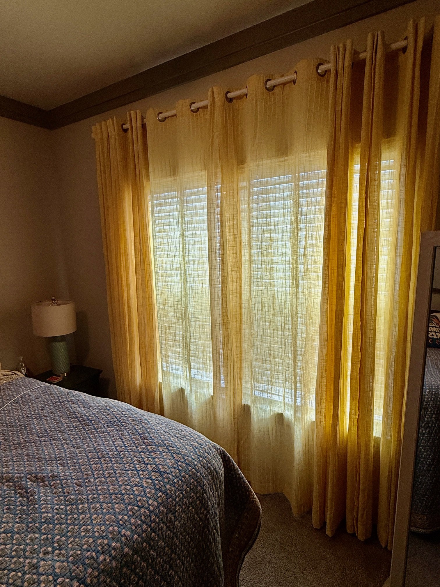 A bedroom corner with yellow curtains hanging over a window, a section of a bed with a patterned quilt, and a nightstand with a lamp.
