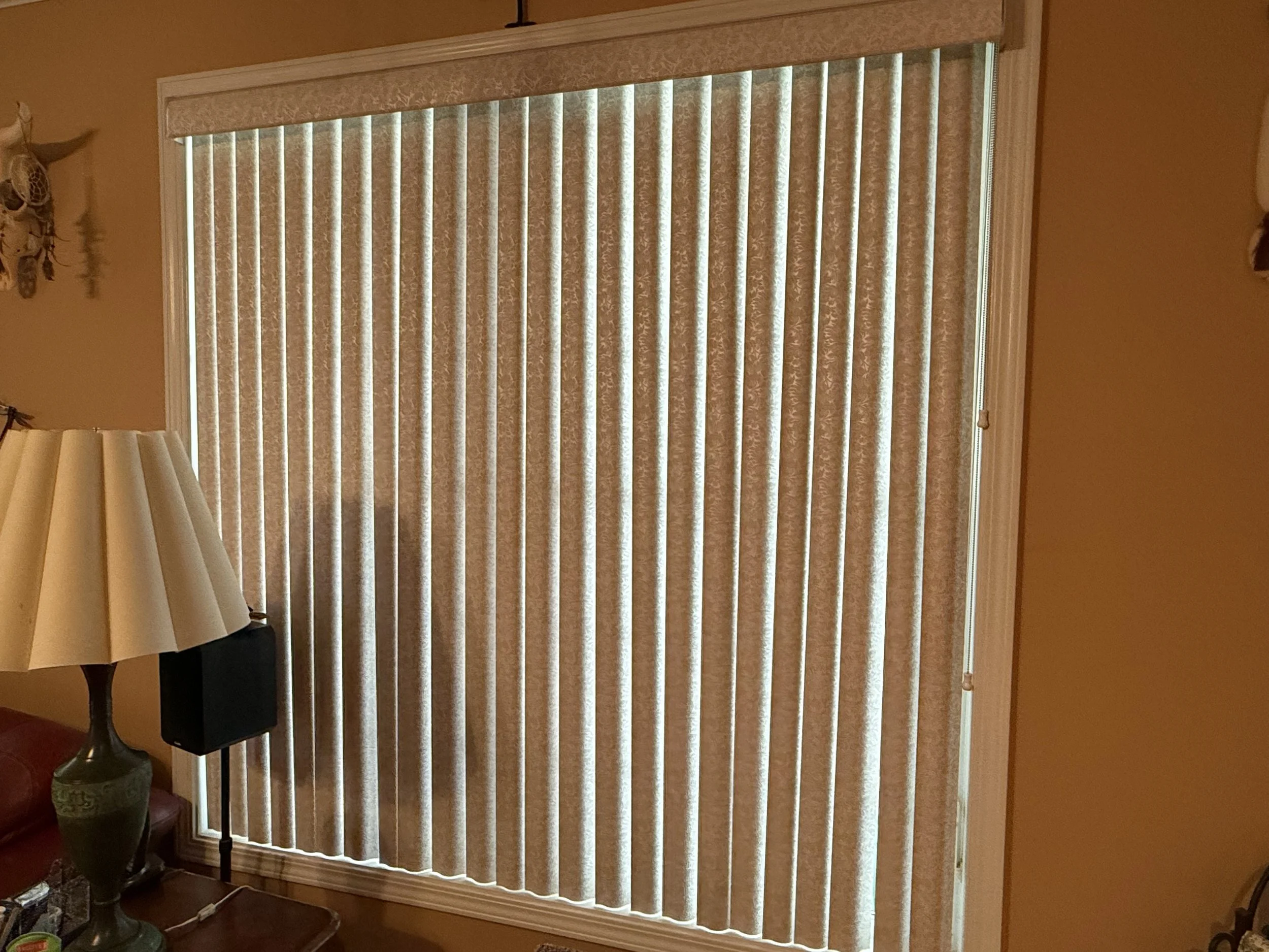 A window with vertical blinds in a room with yellow walls, partially covered by a wooden valance, and a lamp on a table nearby.