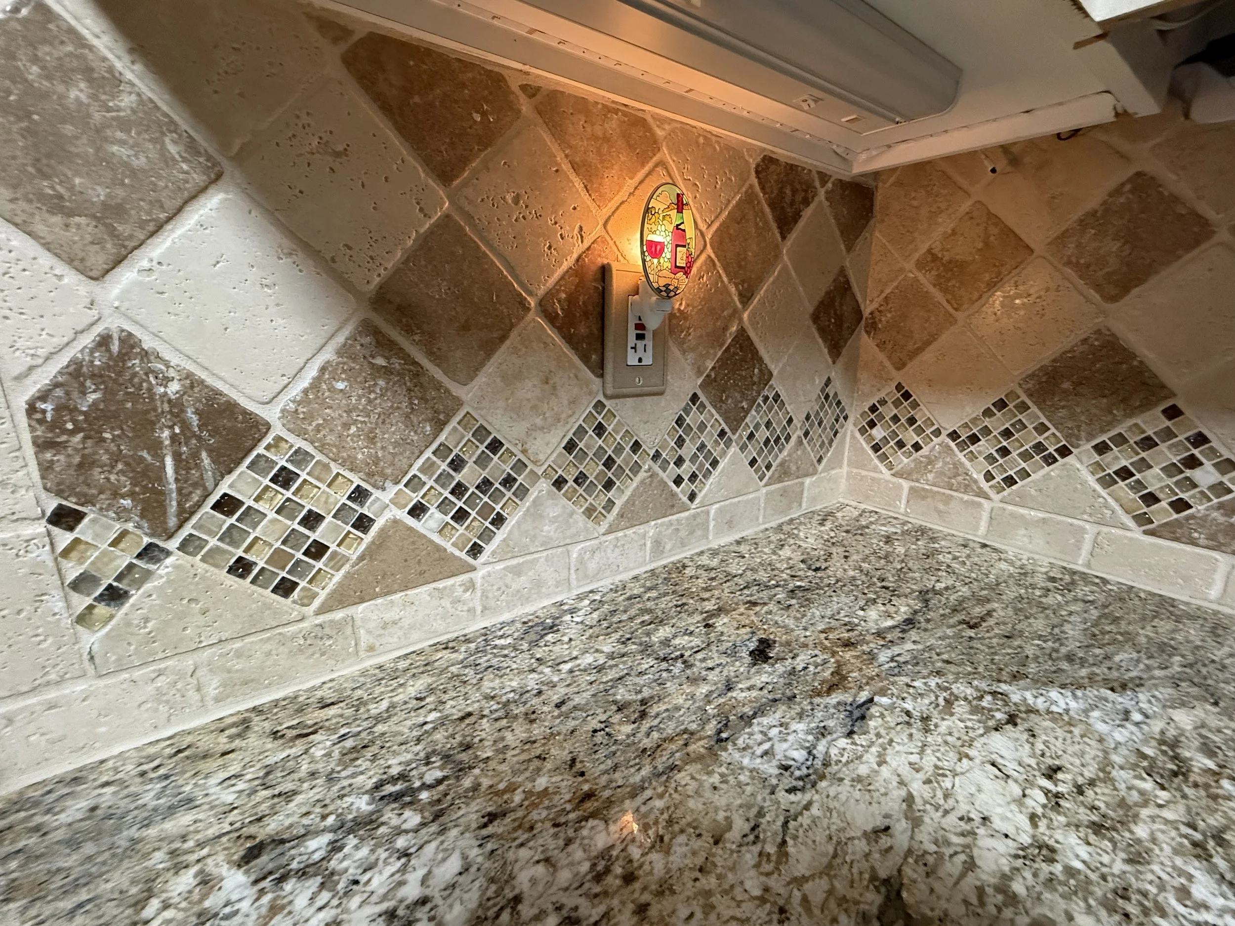 Close-up of a kitchen or bathroom countertop and backsplash. The countertop is made of granite with brown, black, and gray speckles. The backsplash features a combination of large beige and brown stone tiles with some small square mosaic tiles in shades of beige, brown, and black. There is a power outlet with a decorative cover illuminated by a small, warm light.