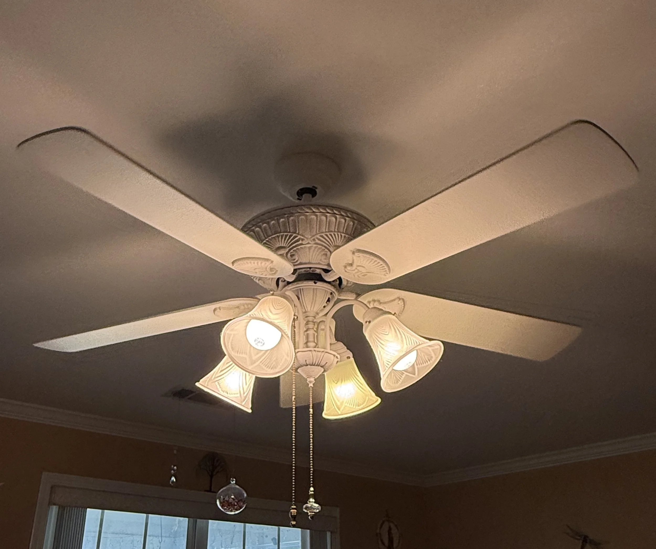 Ceiling chandelier light fixture with multiple cream-colored, petal-shaped lampshades in a room with a window and dark ceiling.