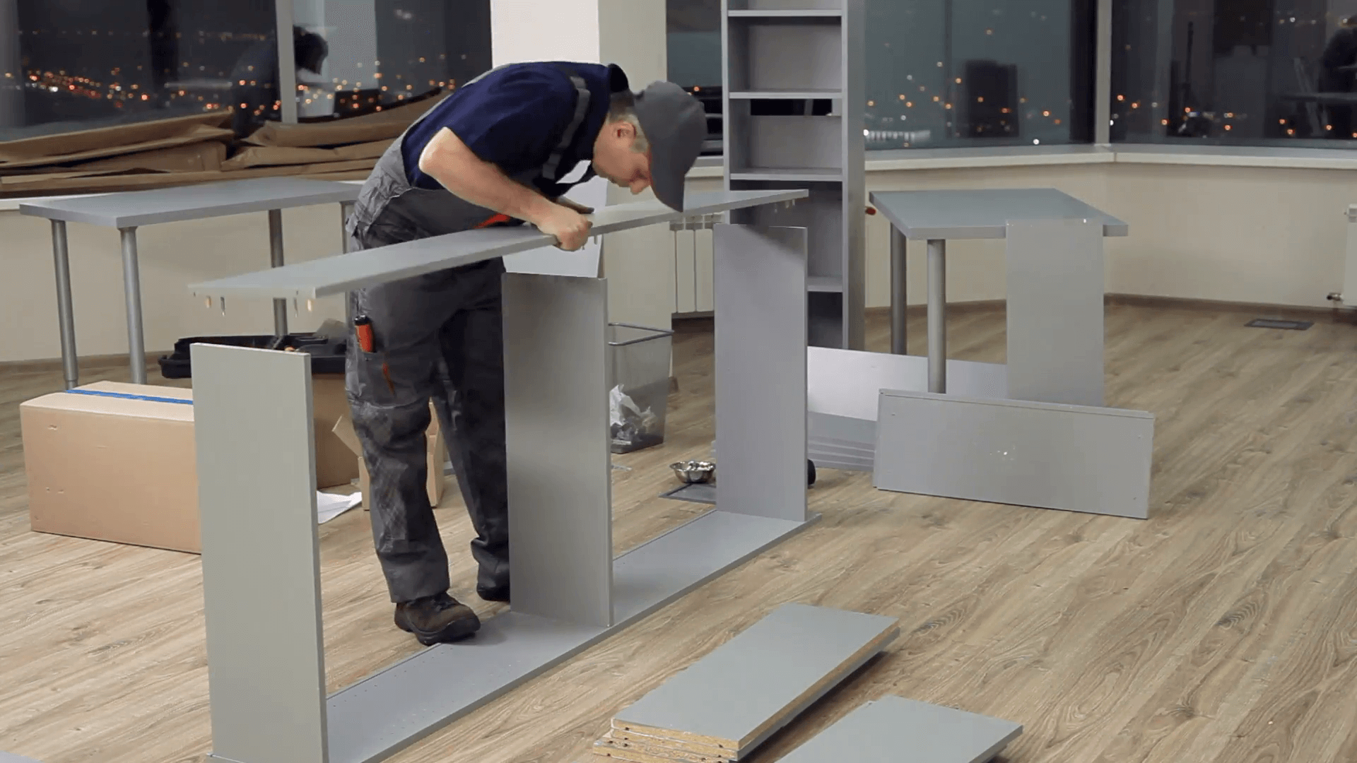 A person assembling furniture in a room with large windows and city lights outside at night.