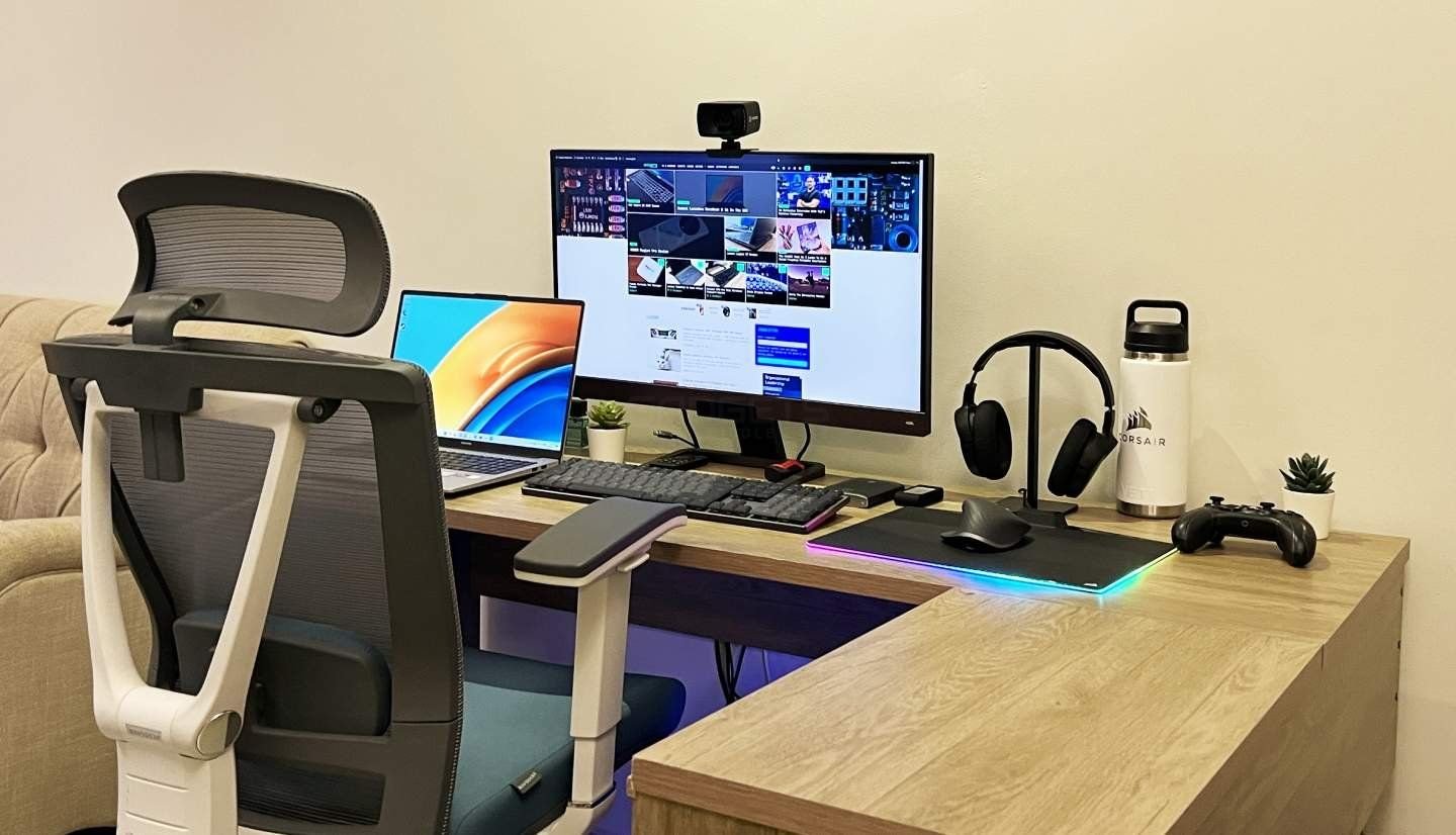 A modern office desk setup with a mesh ergonomic office chair, a laptop with a colorful screen, a large computer monitor displaying a social media feed, a wireless keyboard and mouse, a headset on a stand, a gaming controller, a water bottle, and two small potted plants.
