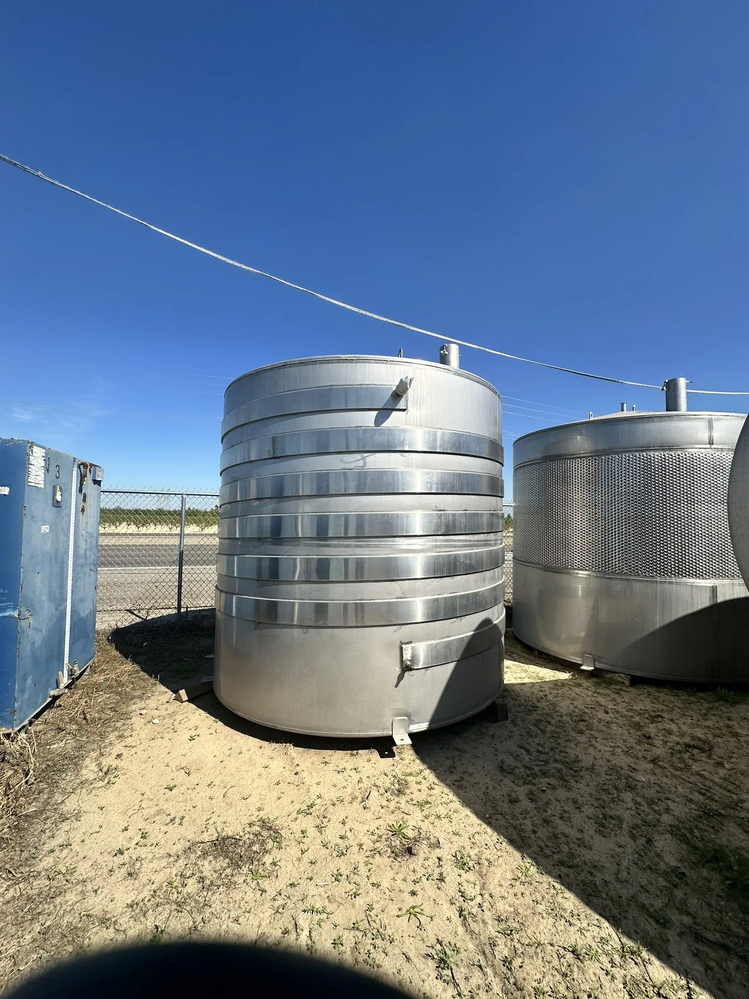 Large cylindrical metal water storage tanks outdoors on a dirt ground, with a chain-link fence and a clear blue sky in the background.