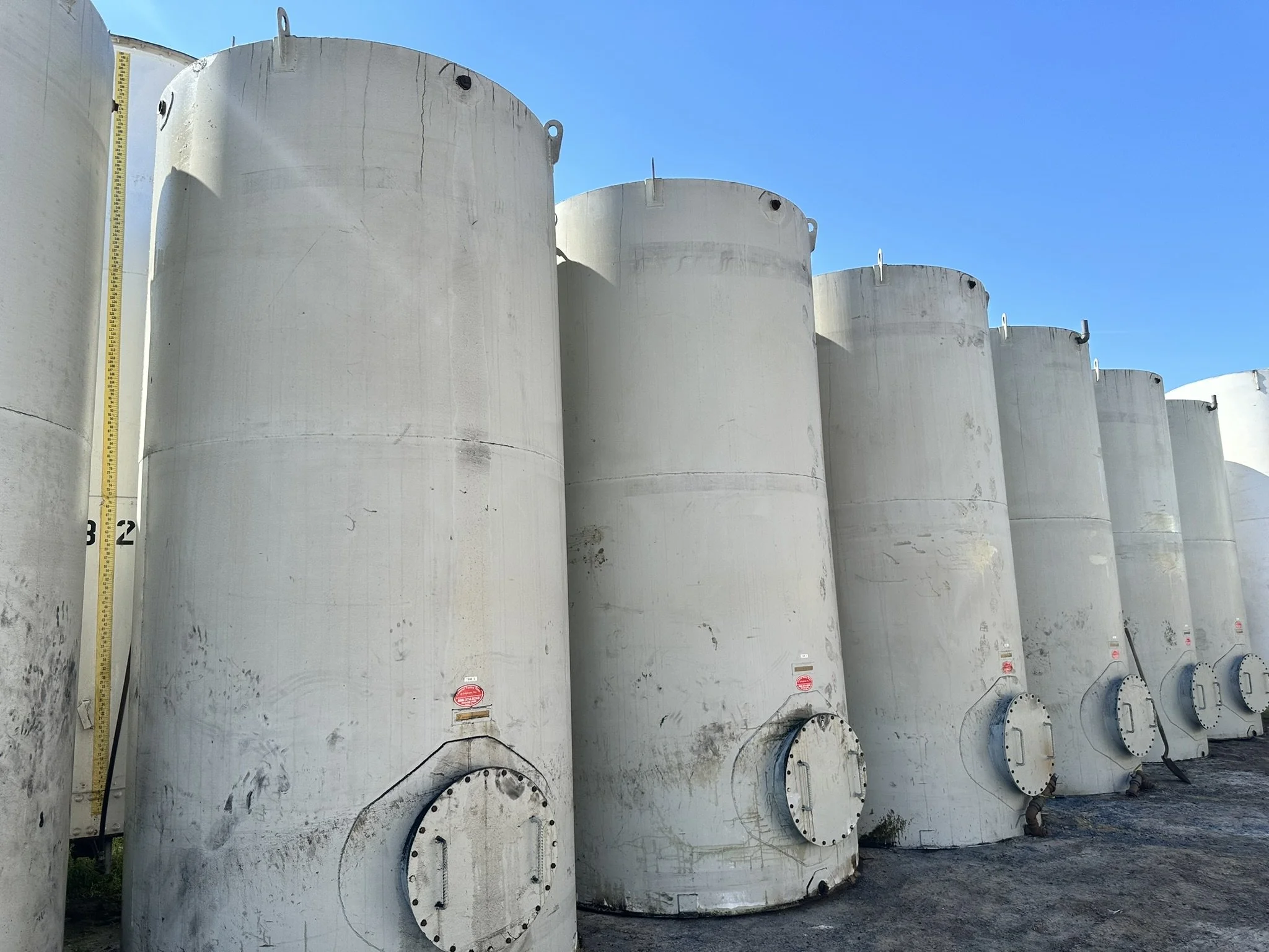 A row of large white industrial storage tanks outdoors under a clear blue sky, with a height measurement mark on one tank.