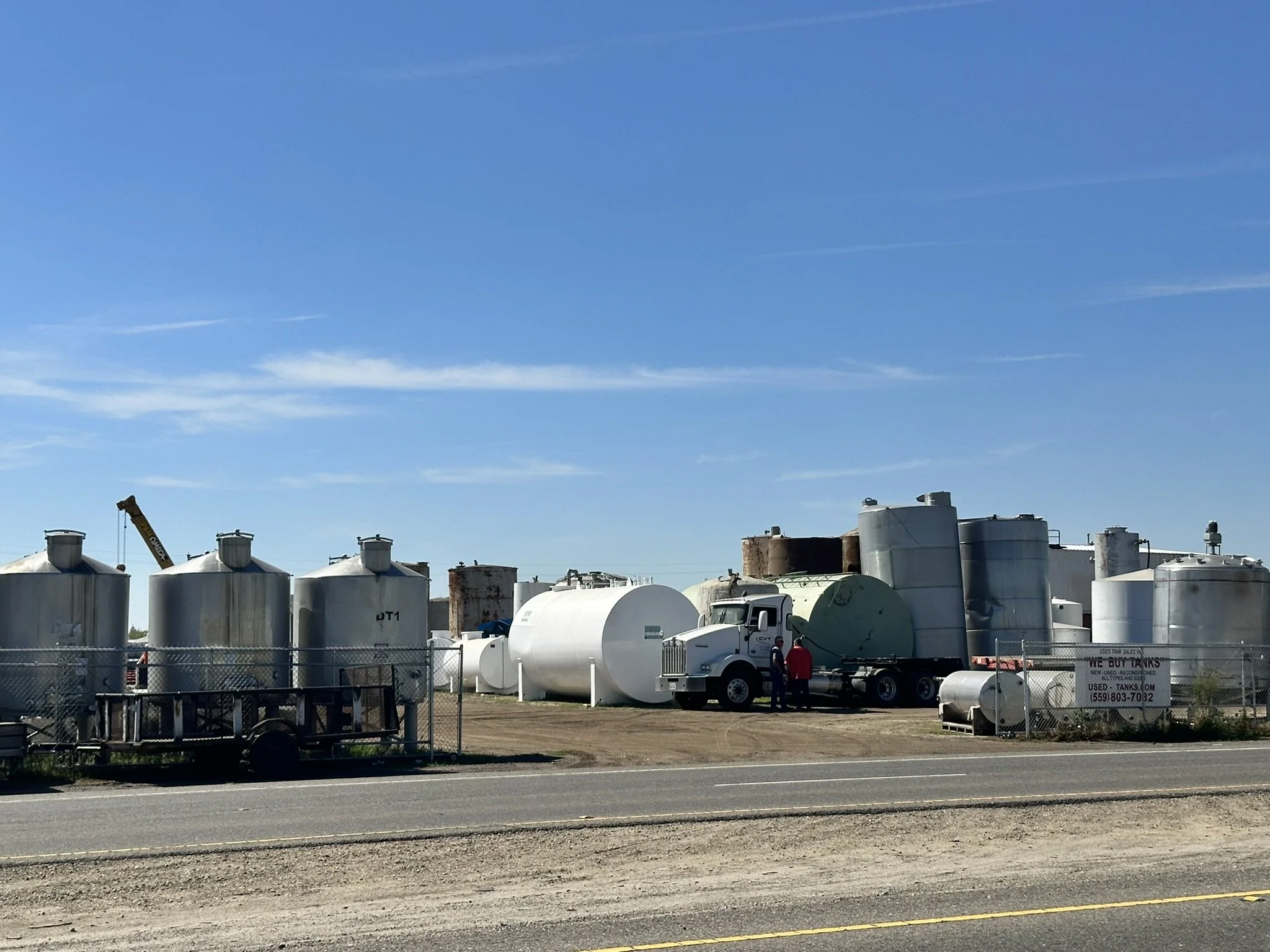 Industrial fuel or chemical storage tanks in an outdoor lot, with a truck and people nearby, under a blue sky.