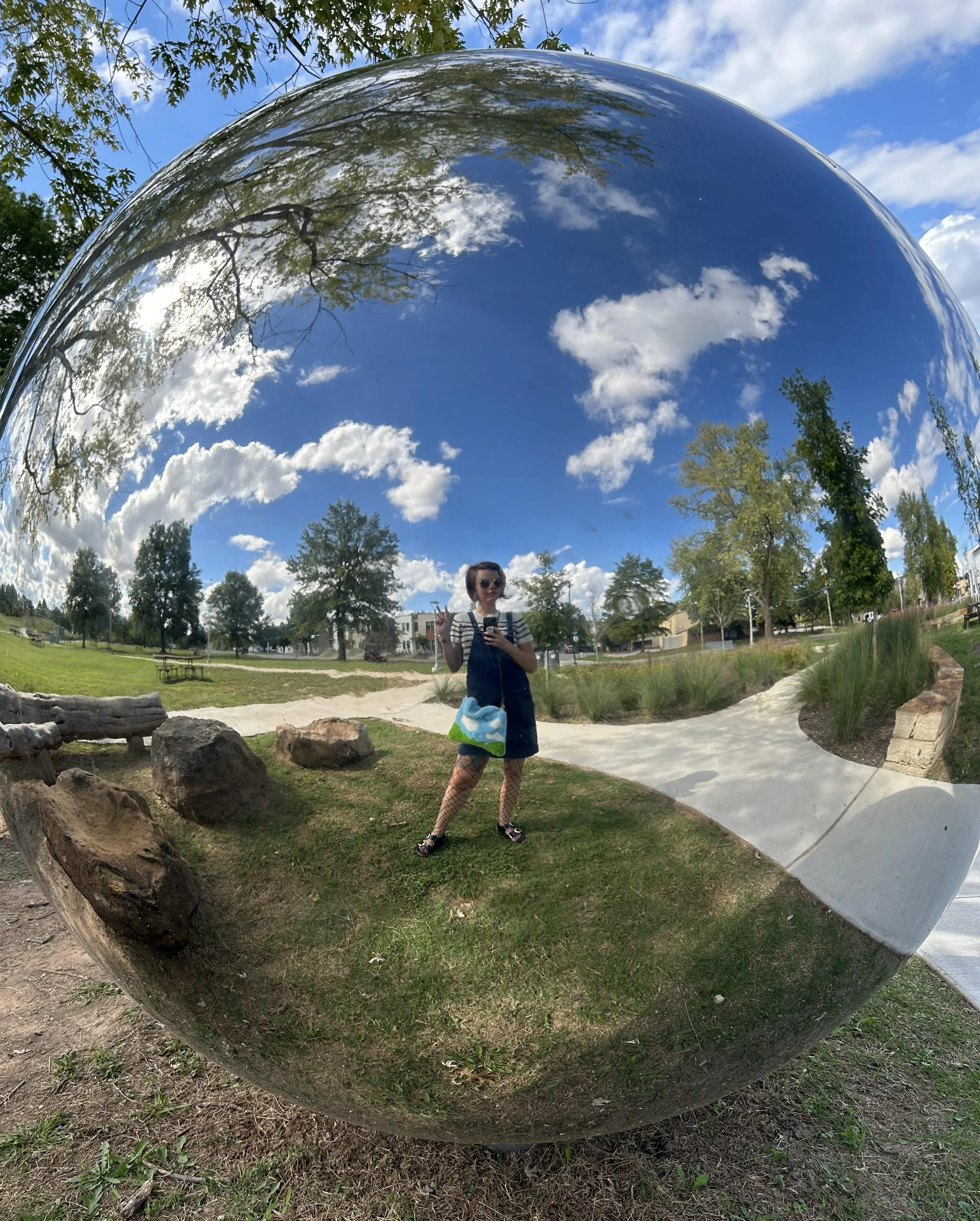 Person taking a photo in a park, reflected in a large reflective spherical sculpture, with trees, sky, and clouds visible.