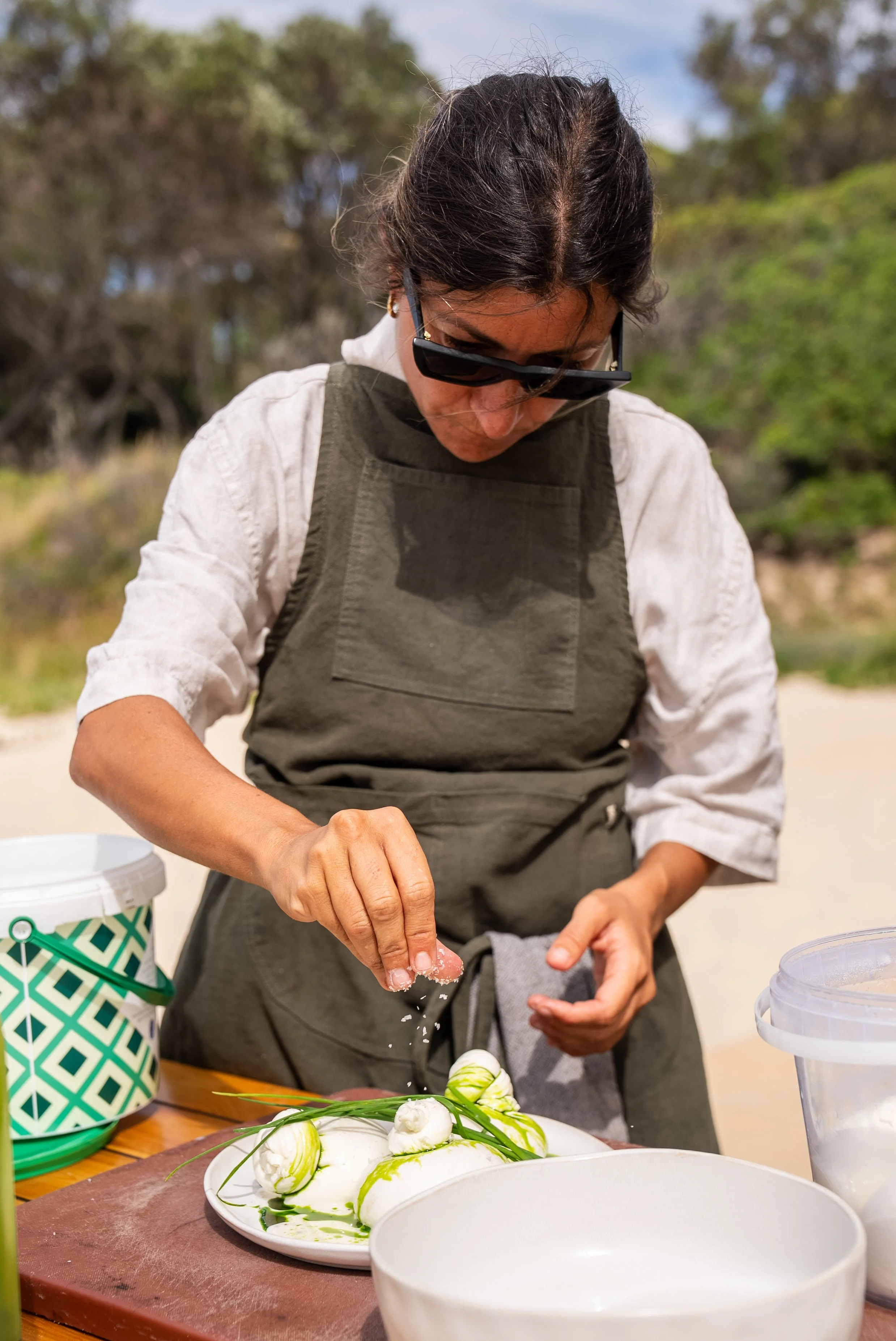 A person wearing sunglasses and a gray apron is preparing food outdoors, with a plate of mozzarella balls and basil on a cutting board, and a white bowl nearby.