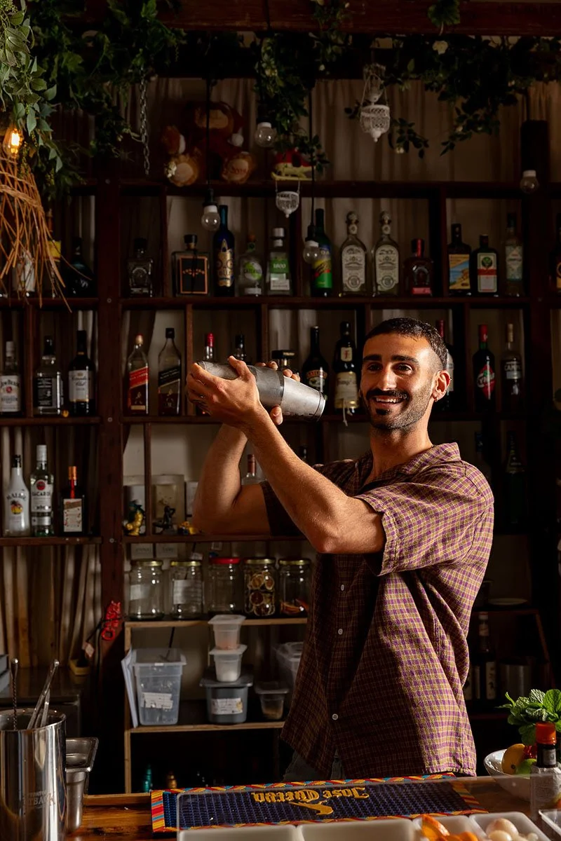 A bartender preparing a drink behind a bar with shelves of liquor bottles in the background.