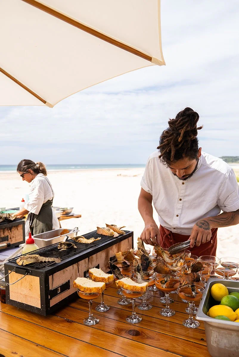 Two chefs preparing seafood dishes on a beachside setup under a large white umbrella, with the ocean and sky in the background.