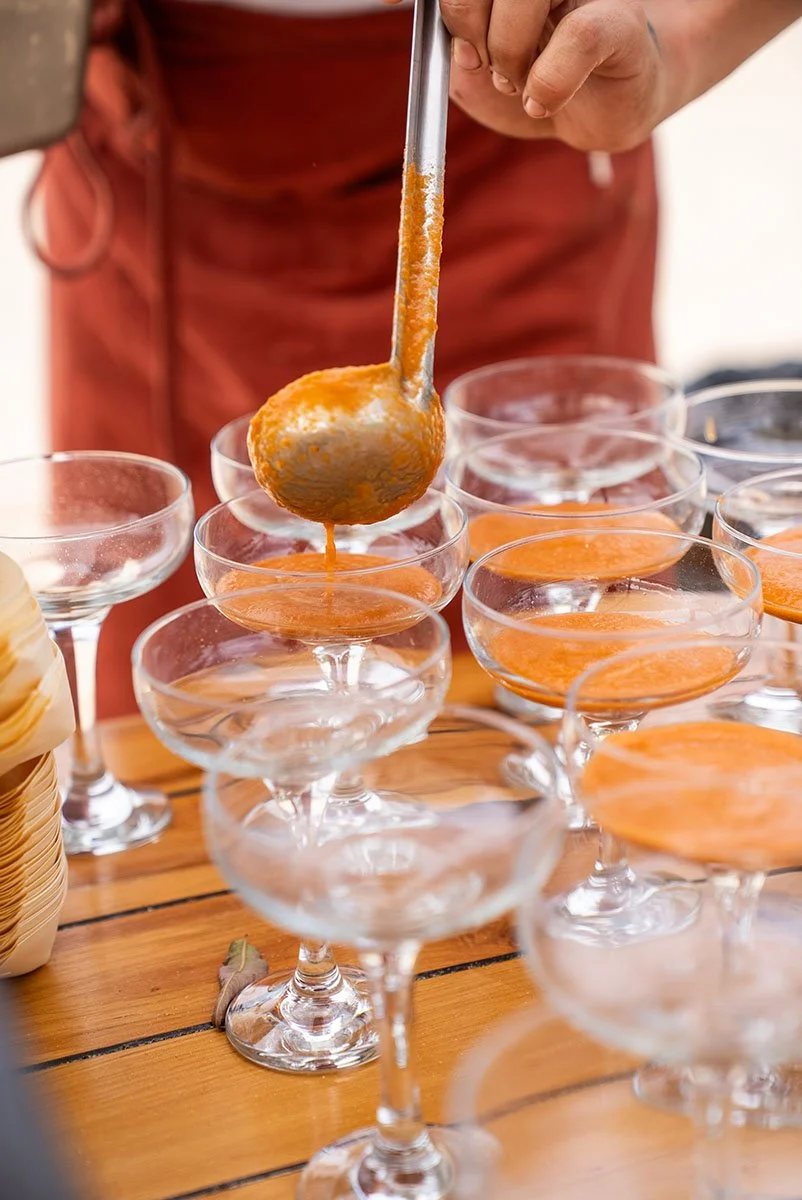 Person serving orange-colored drink into margarita glasses on a wooden table.