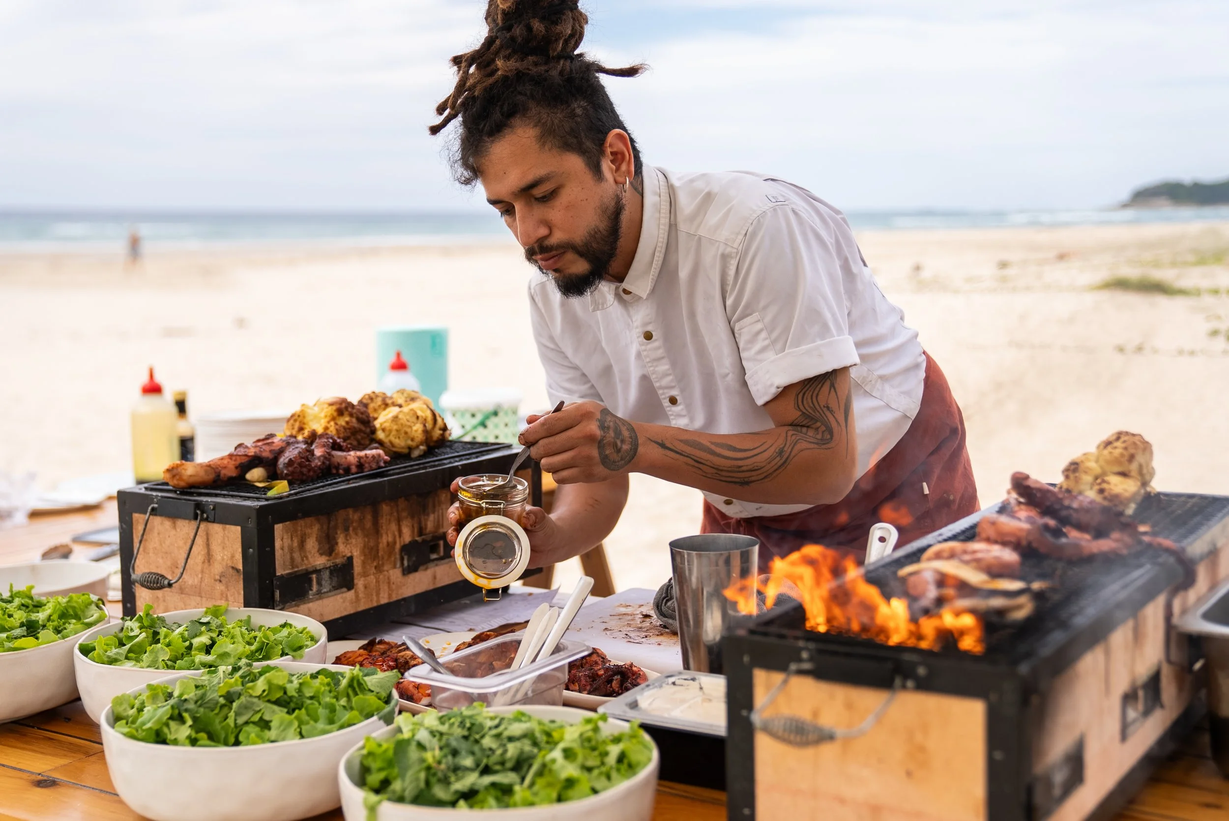 A man with dreadlocks and tattoos grilling meat and preparing food at a beachside outdoor barbecue with a table of salads and condiments.