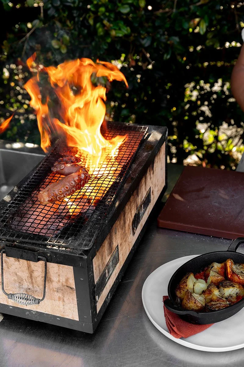 Grilling octopus tentacles over an open flame on a portable outdoor grill, with roasted vegetables in a cast iron skillet nearby on a white plate.