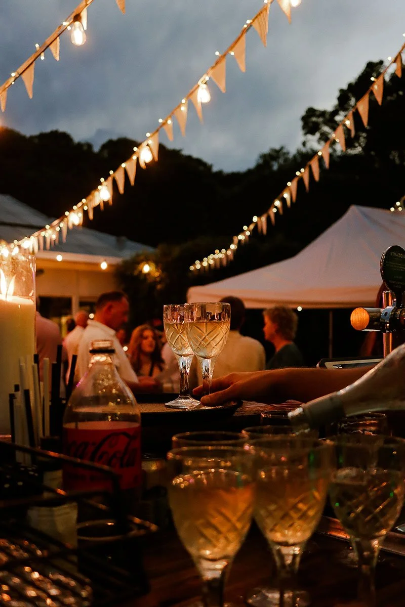 An outdoor gathering at dusk with string lights and tents, people socializing, and glasses of drinks on a bar counter.