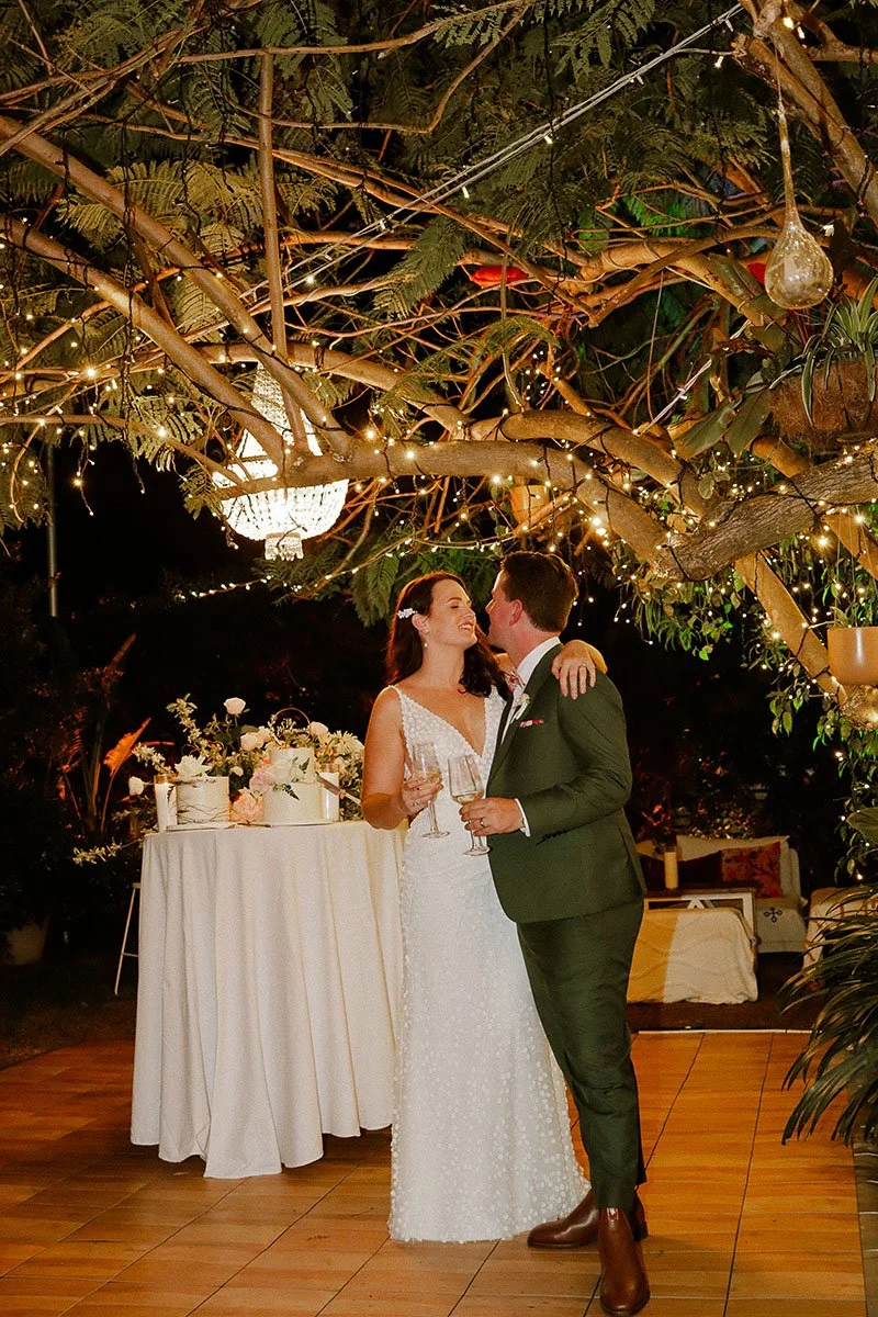 A bride and groom at their wedding reception, holding glasses of champagne under a canopy of tree branches decorated with string lights and a chandelier.