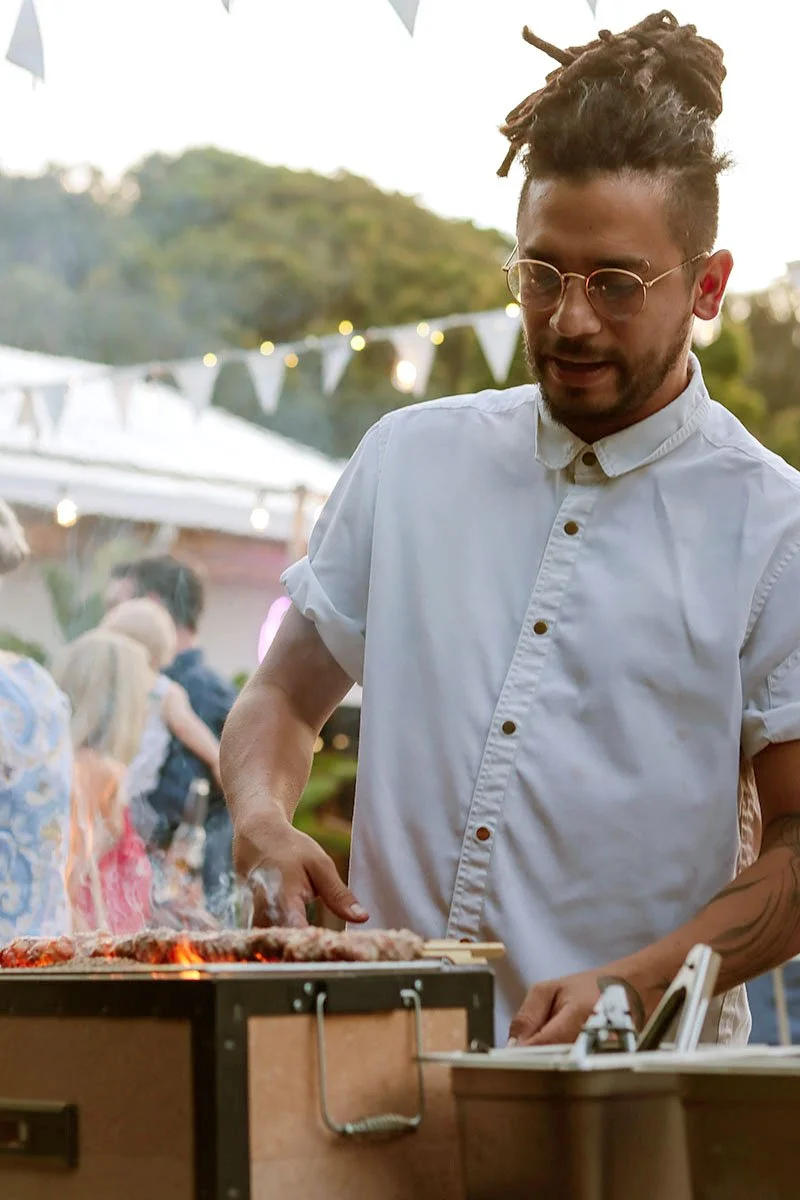 Man with dreadlocks wearing sunglasses and a white button-up shirt grilling meat at an outdoor event with string lights and people in the background.