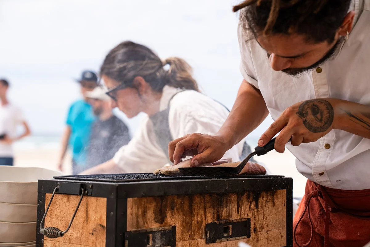 A man grilling fish on an outdoor barbecue at a beach, with other people in the background.