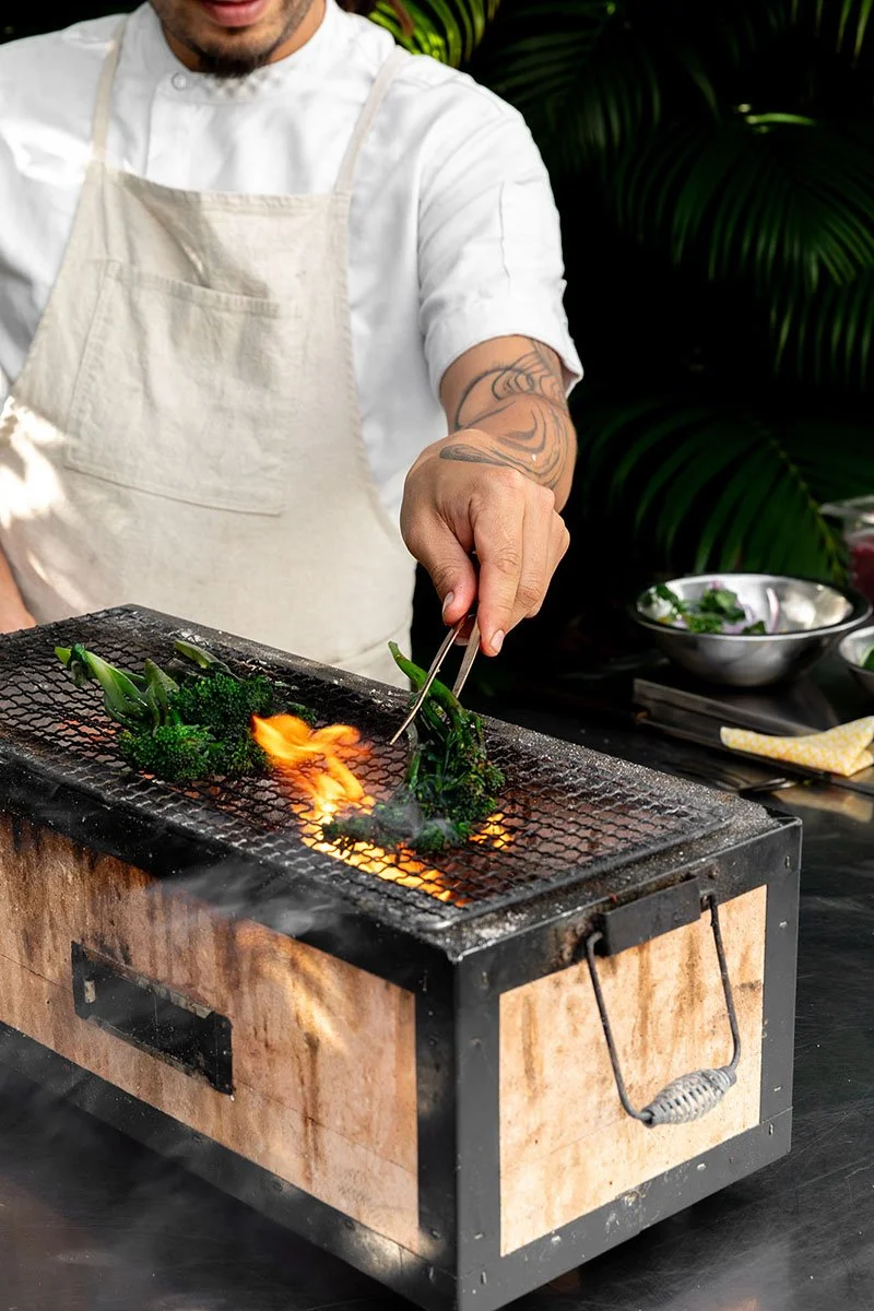 A chef wearing a white apron and shirt uses tongs to handle green vegetables on a small tabletop grill with flames. The background includes leafy plants and metal bowls on a black surface.