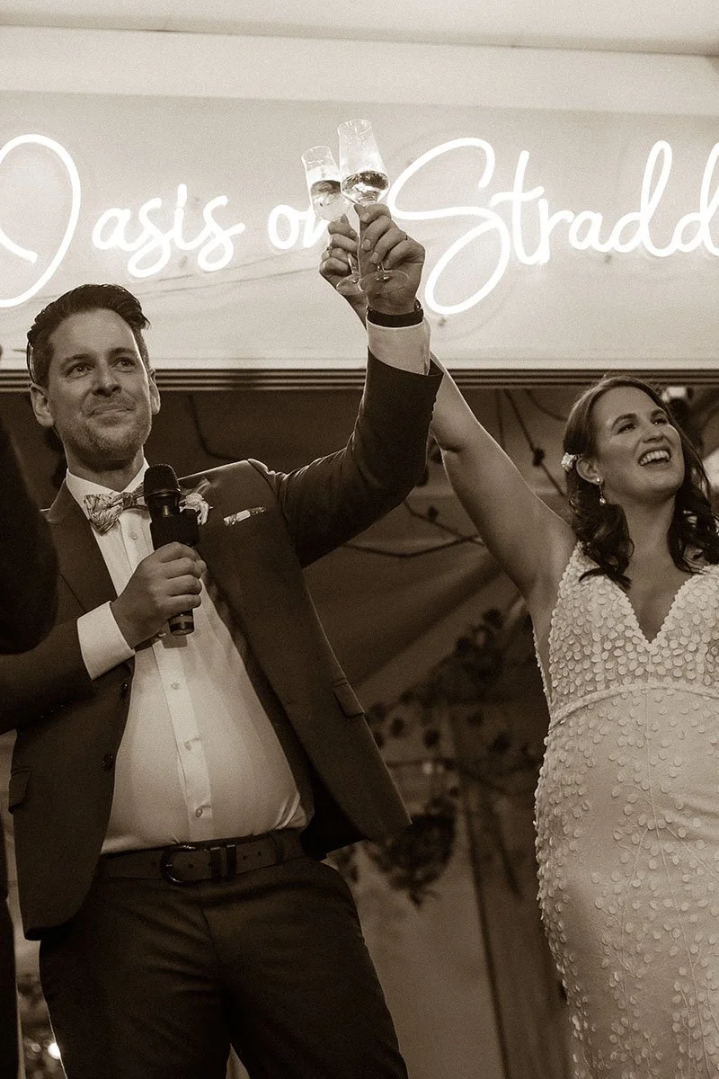 A man and woman in wedding attire at a celebration, raising champagne glasses in a toast. The man is wearing a tuxedo with a bow tie, and the woman is in a white wedding dress. They are smiling and standing in front of a sign that reads 'Oasis of Str