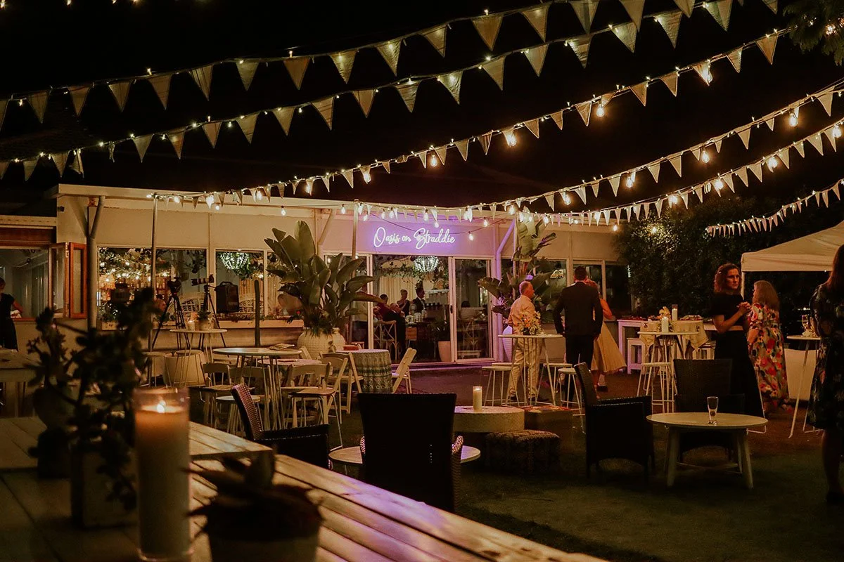 Nighttime outdoor party with string lights and triangular pennant banners hanging above. People are standing and sitting around tables, some chatting. A tent and a building with a neon sign reading 'Oasis on the Straddle' are visible in the backgroun