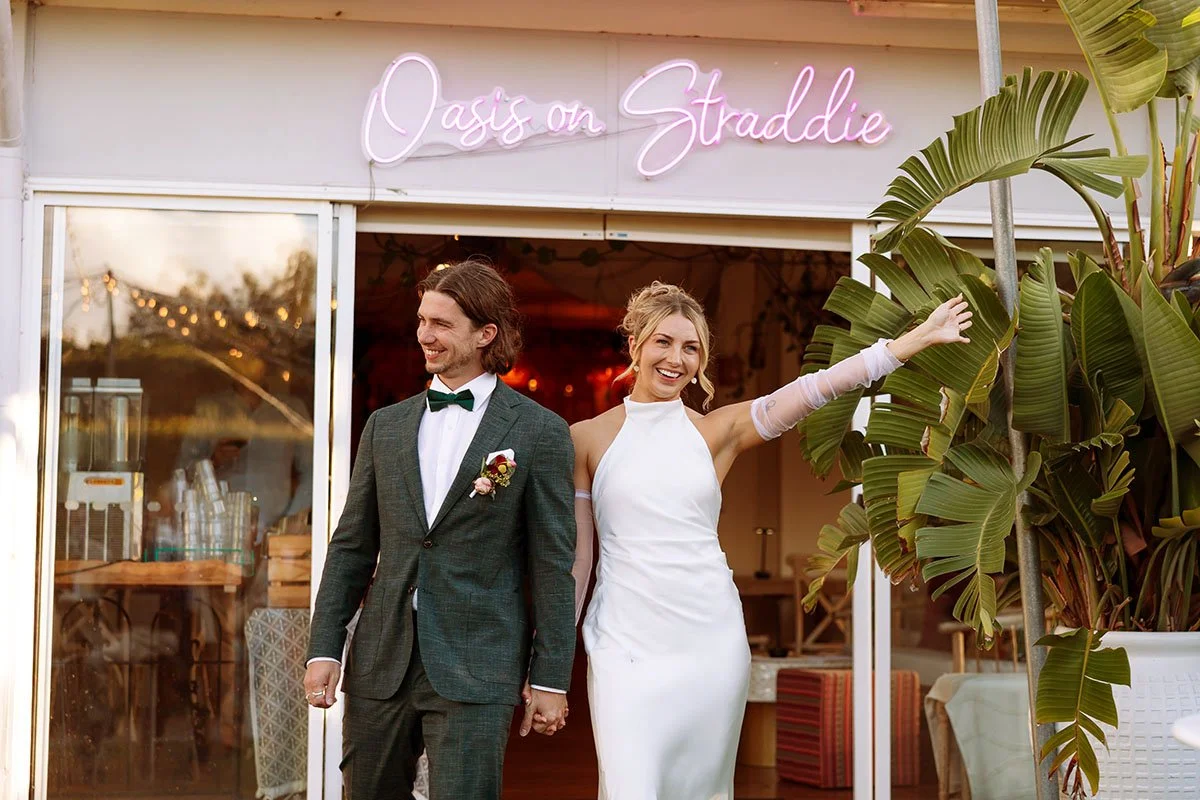 A bride and groom standing hand in hand outside a venue with a neon sign that reads 'Oasis on Straddie.' The bride is smiling and touching a large potted plant, while the groom is looking at her. The setting appears to be a wedding celebration.
