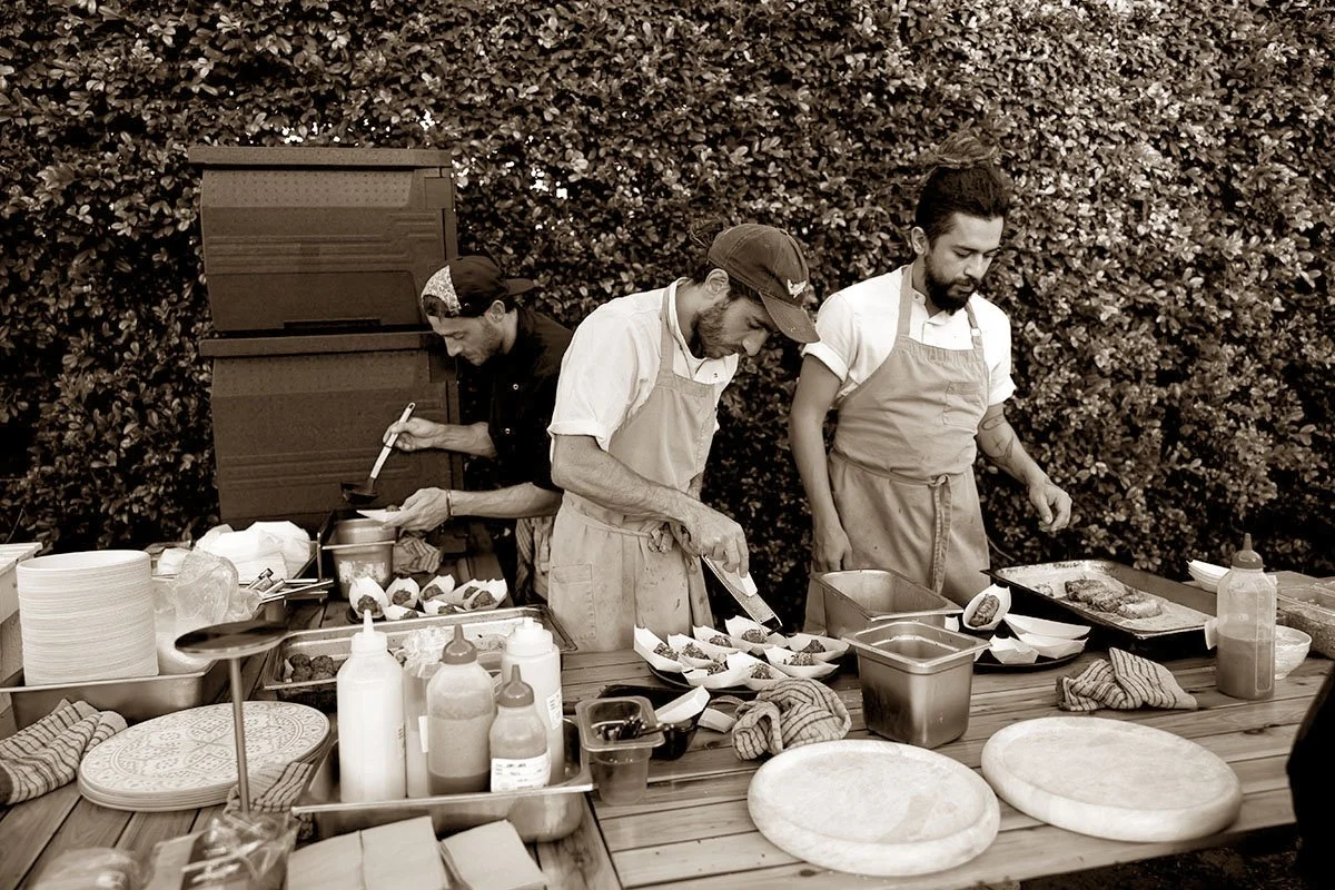 Three chefs preparing food on a wooden outdoor table with plates, condiments, and kitchen tools, surrounded by a hedge.