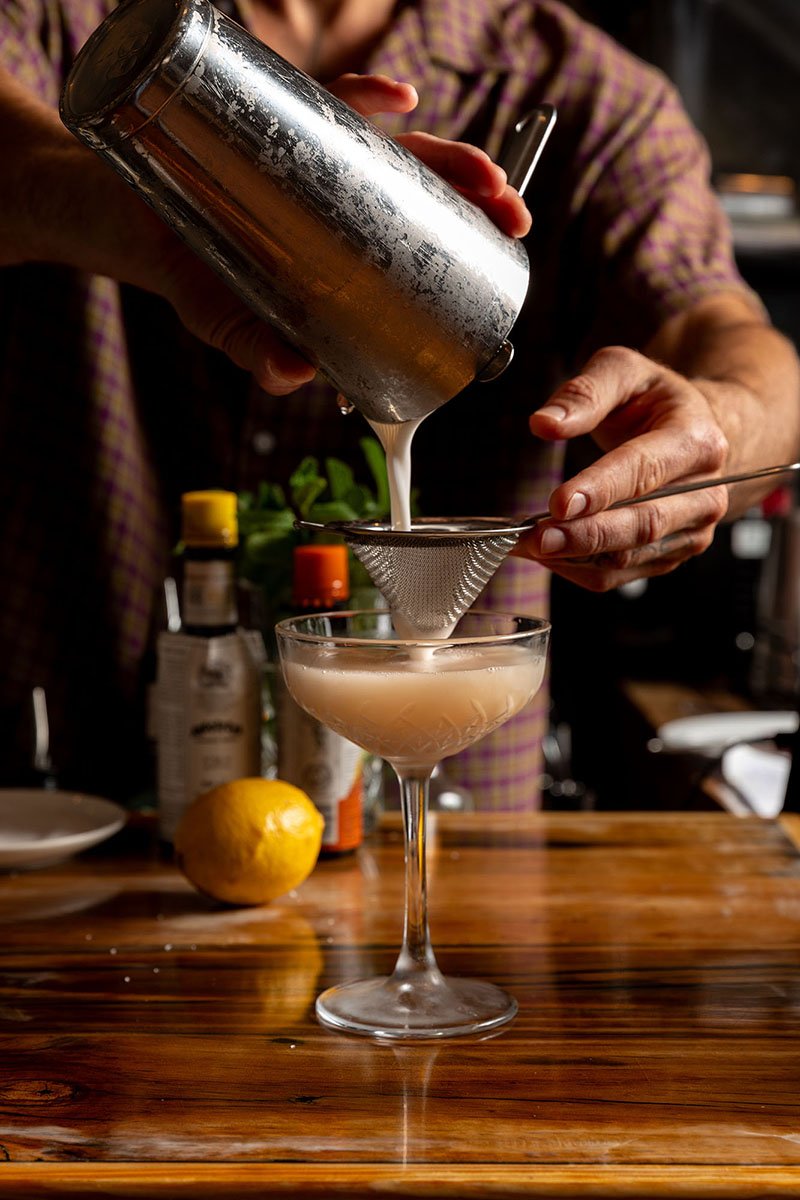 A bartender pours a creamy cocktail through a strainer into a coupe glass, with a lemon and bottles on a wooden bar behind.