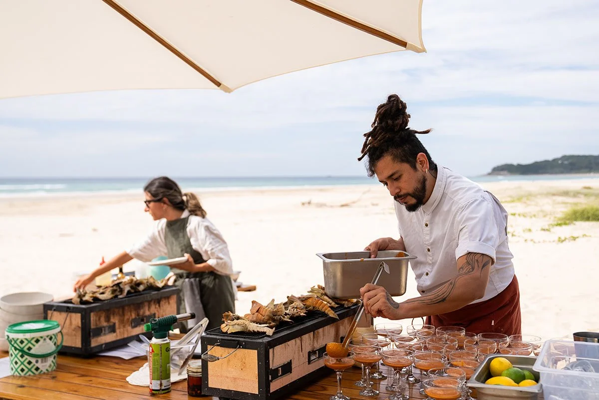 A man with dreadlocks serving seafood in glasses at a beachside outdoor event, with a woman preparing food in the background under a large umbrella.