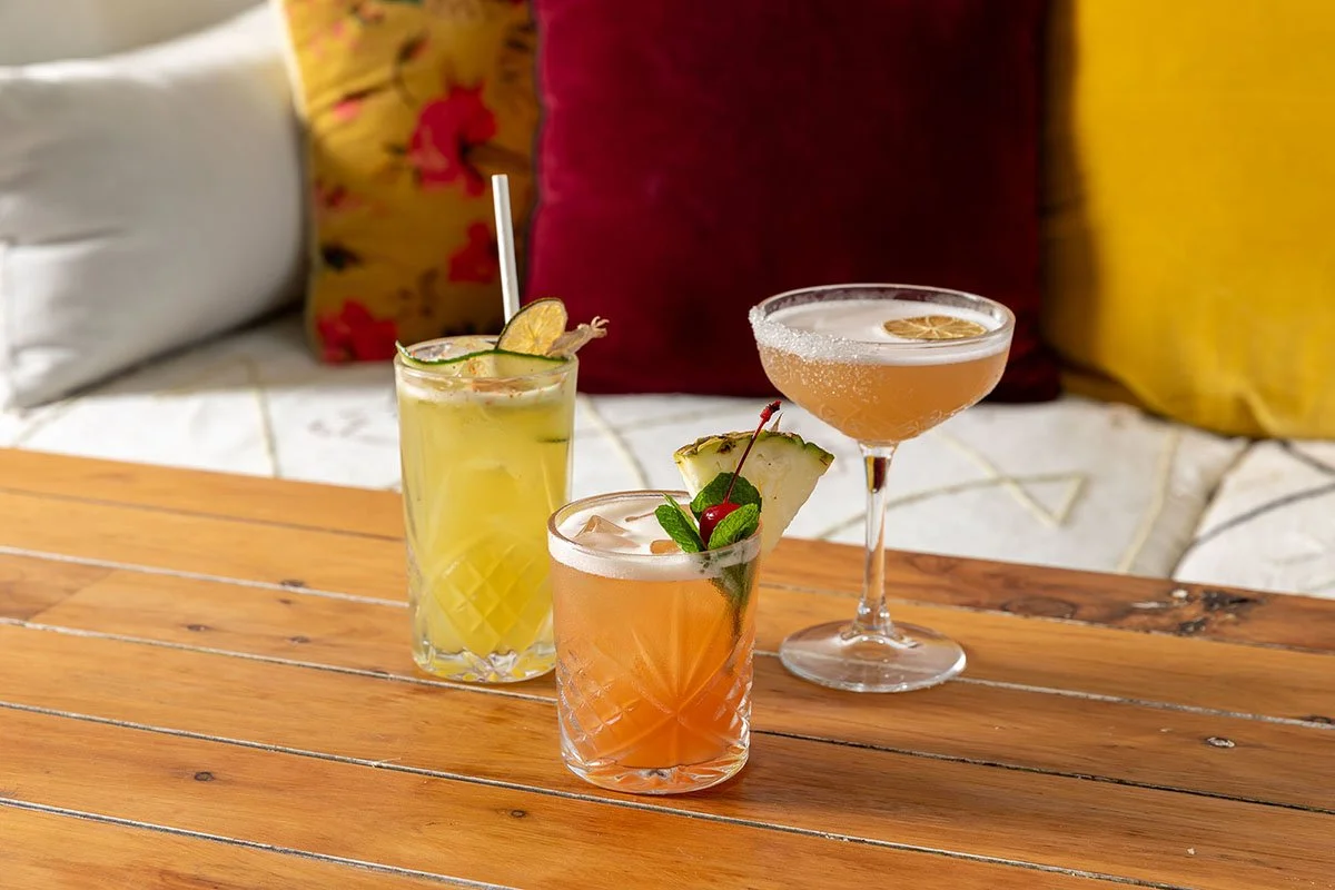 Three colorful cocktails on a wooden table in front of decorative pillows.