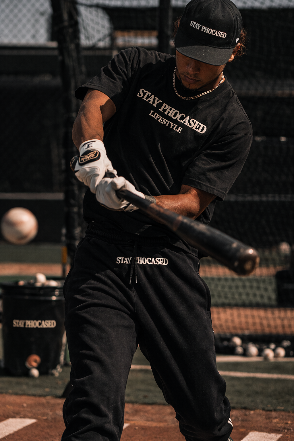 A person practicing baseball swings at a ball on a practice field, wearing black workout clothes with the phrase 'STAY PHOCUSED' printed on them, a black cap, and gloves.