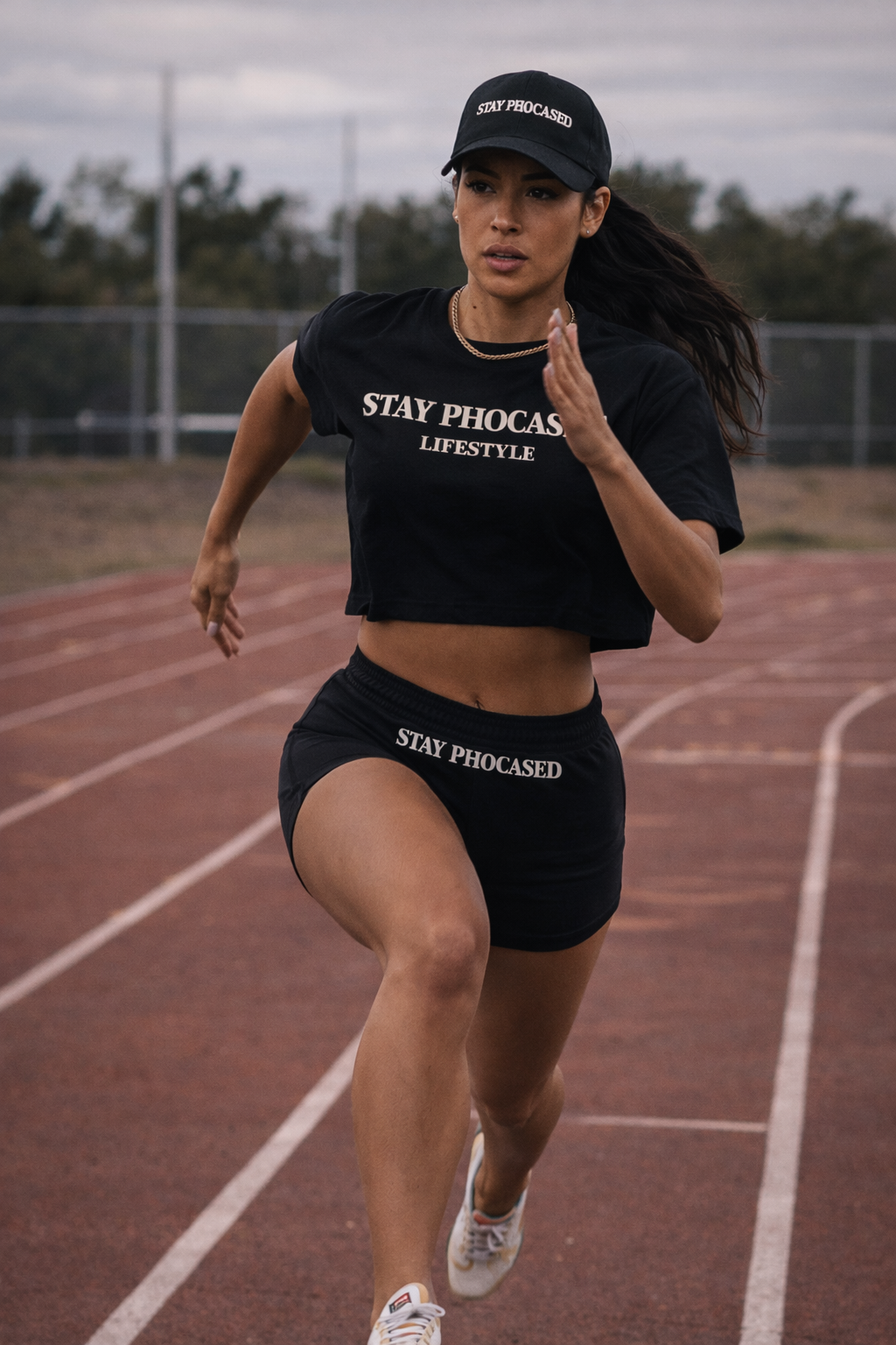 A woman running on a track wearing a black cap, black crop top, and black shorts, all featuring the text 'STAY PHOCASD.'