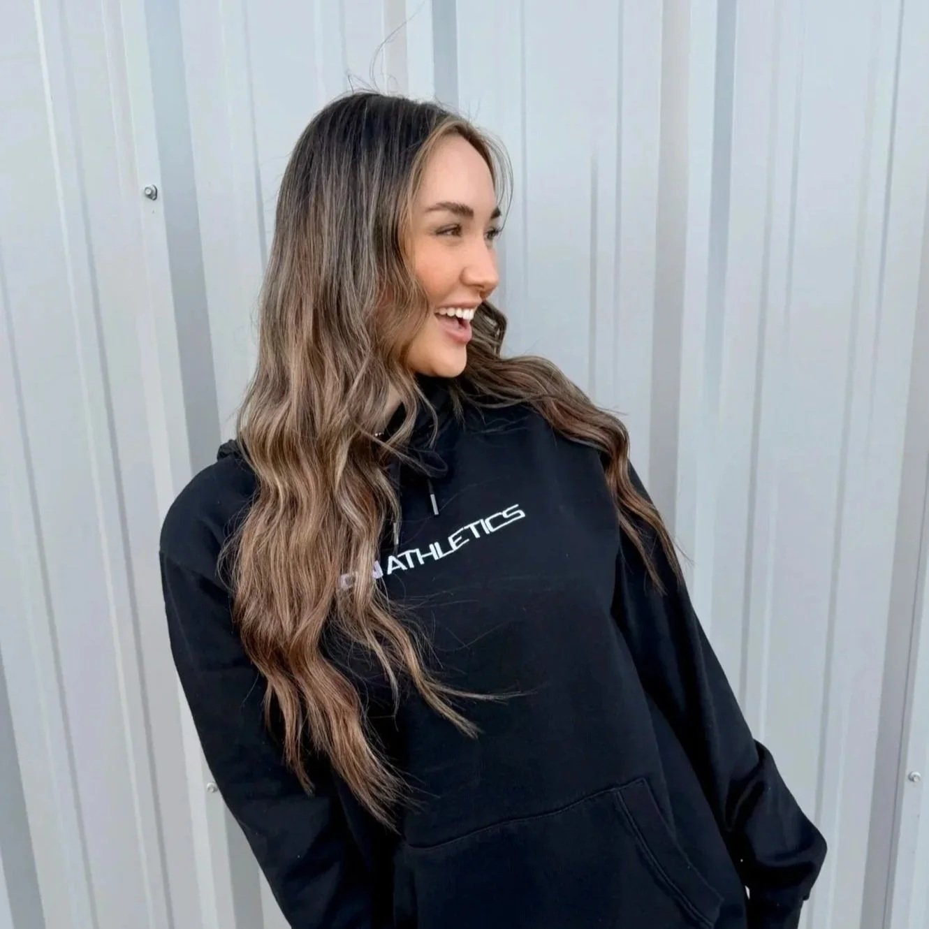 A young woman with long wavy brown hair wearing a black hoodie standing in front of a light gray metal wall smiling and looking to her left.