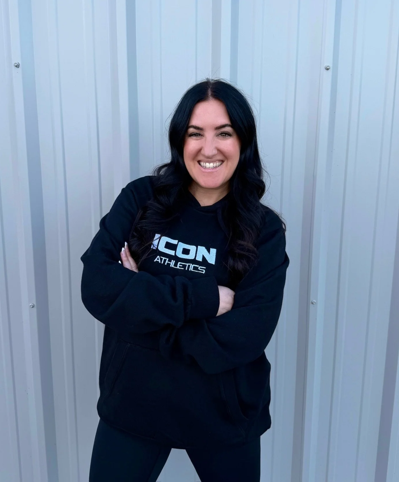 Woman with long dark wavy hair smiling, wearing a black hoodie with 'ICON ATHLETICS' logo, standing in front of a metal wall with arms crossed.