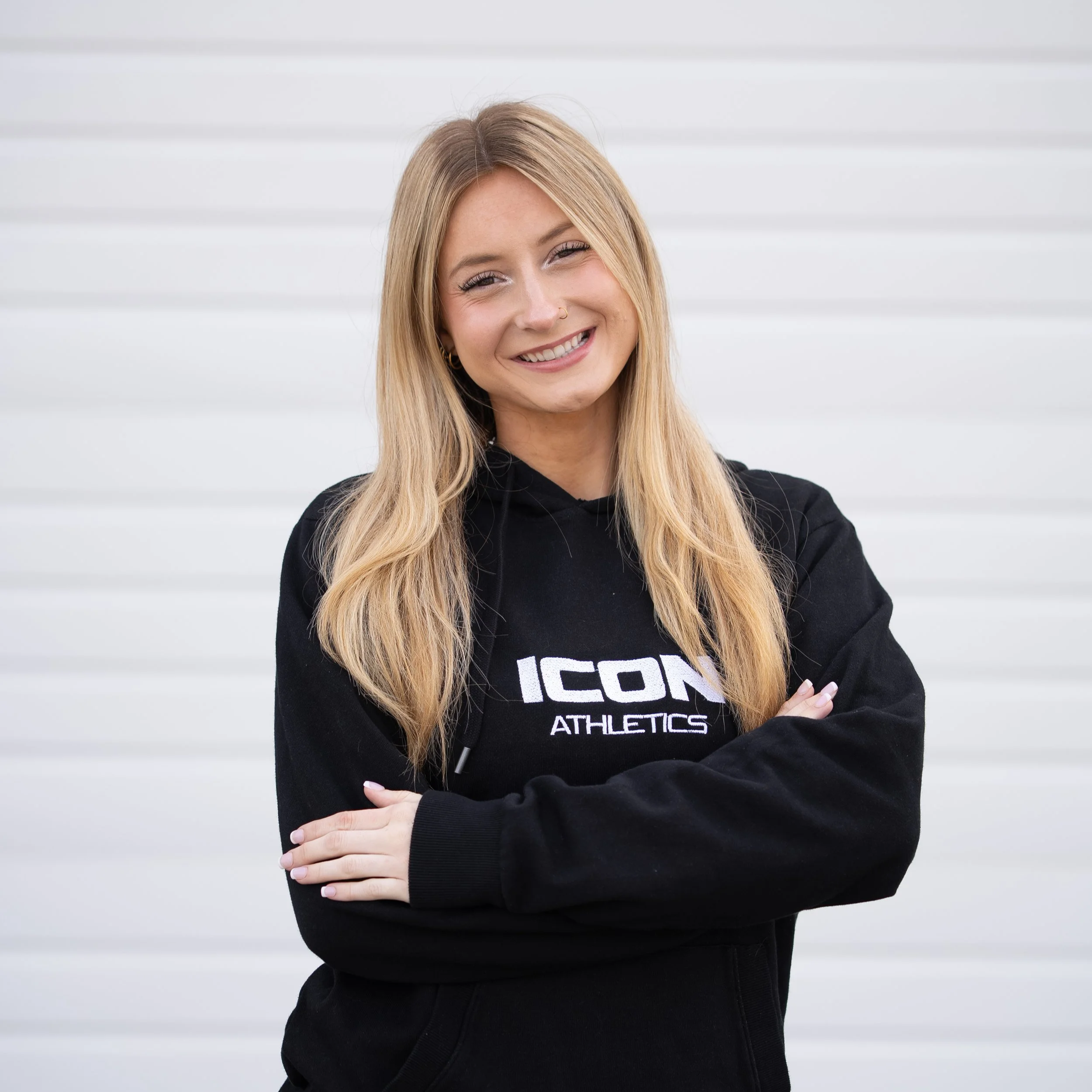 A smiling young woman with long blonde hair wearing a black hoodie with 'ICON ATHLETICS' on it, standing in front of a white background.
