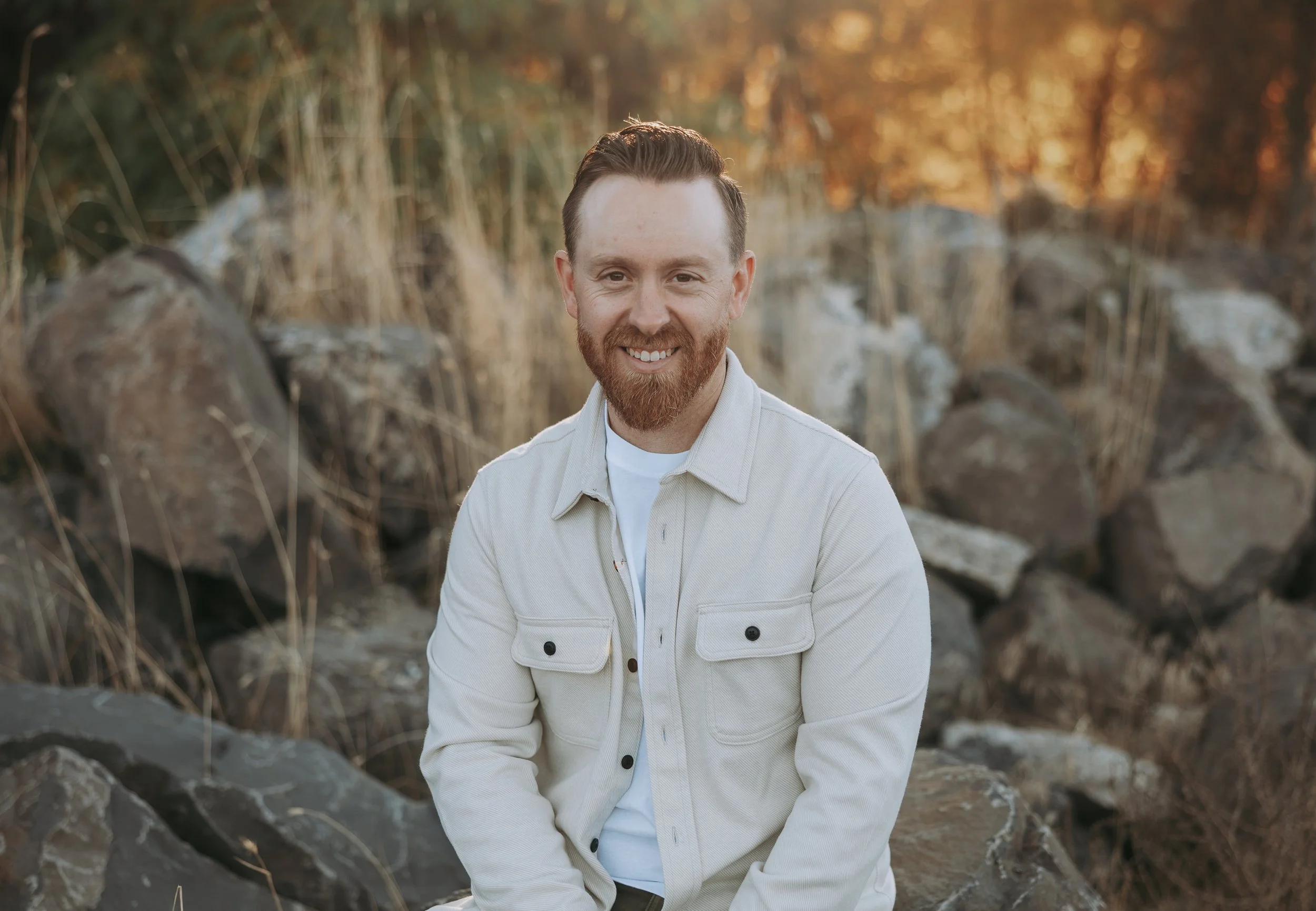 A smiling man with red hair and beard sitting outdoors among rocks and dry grass during sunset, wearing a light-colored jacket and white shirt.