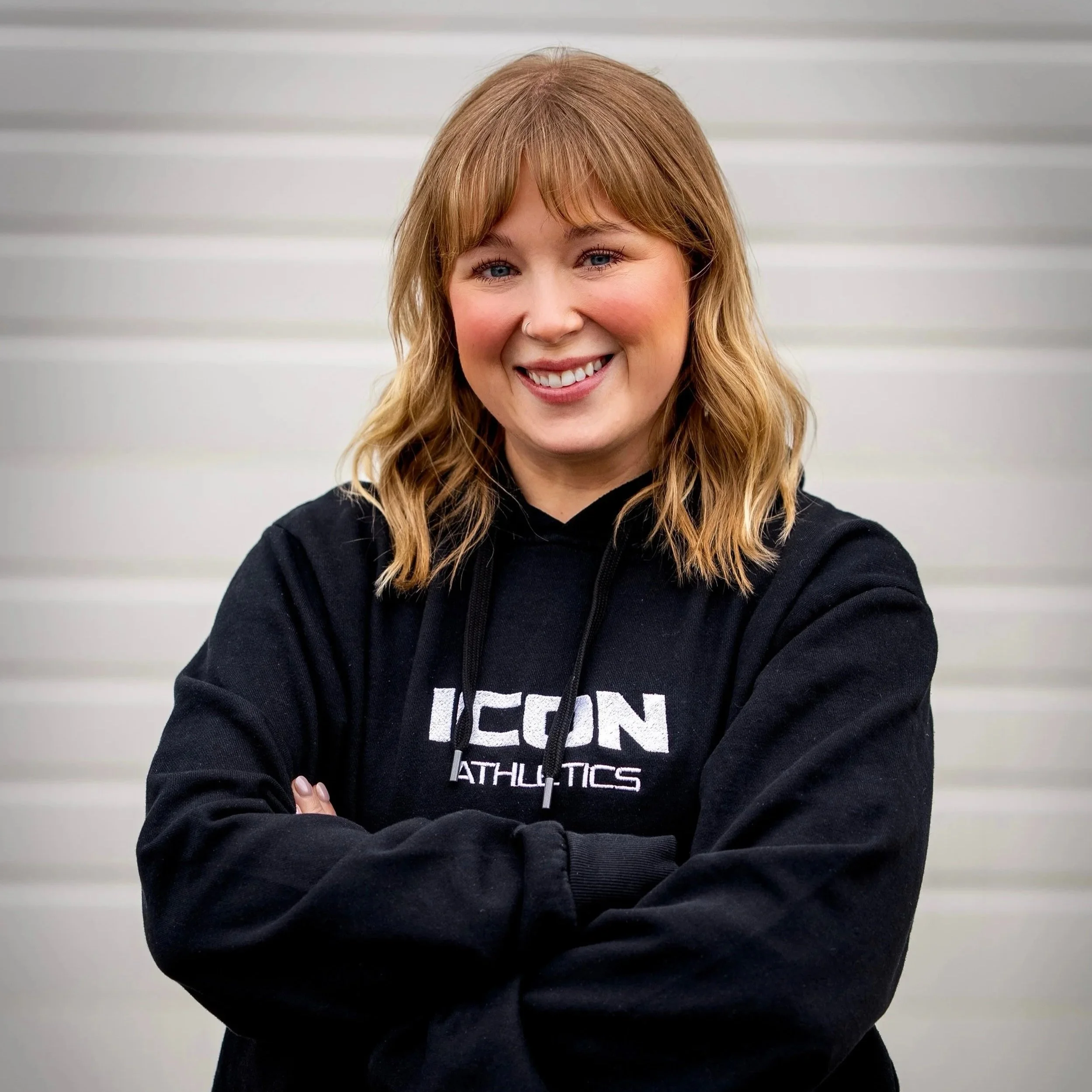 A woman with shoulder-length blonde hair and a nose piercing smiling with arms crossed, wearing a black hoodie with the word "ICON" visible on the front, standing against a light gray horizontal panel wall.