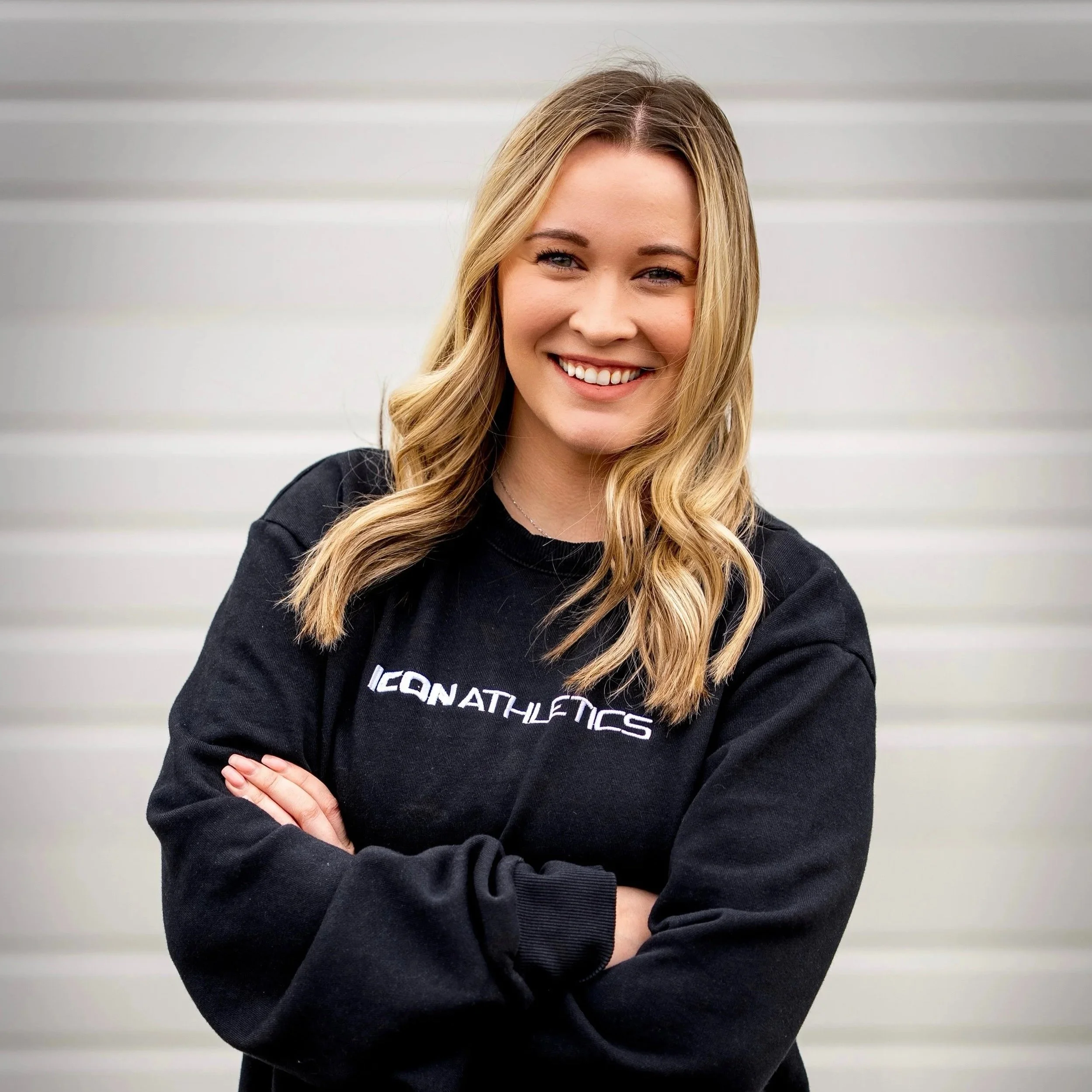 A young woman with blonde hair smiling, wearing a black sweatshirt with white text, standing with her arms crossed in front of a light-colored wall.