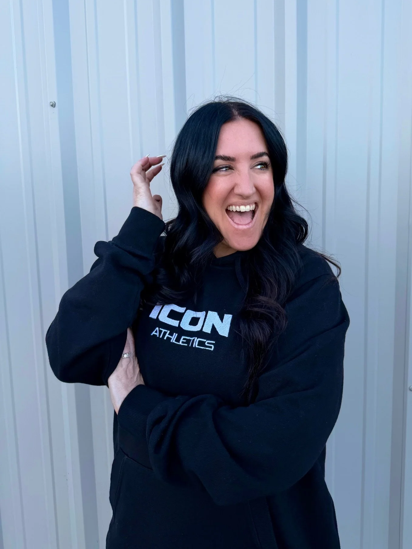 Woman in black ICON Athletics hoodie laughing and playfully touching her hair in front of a metal wall.