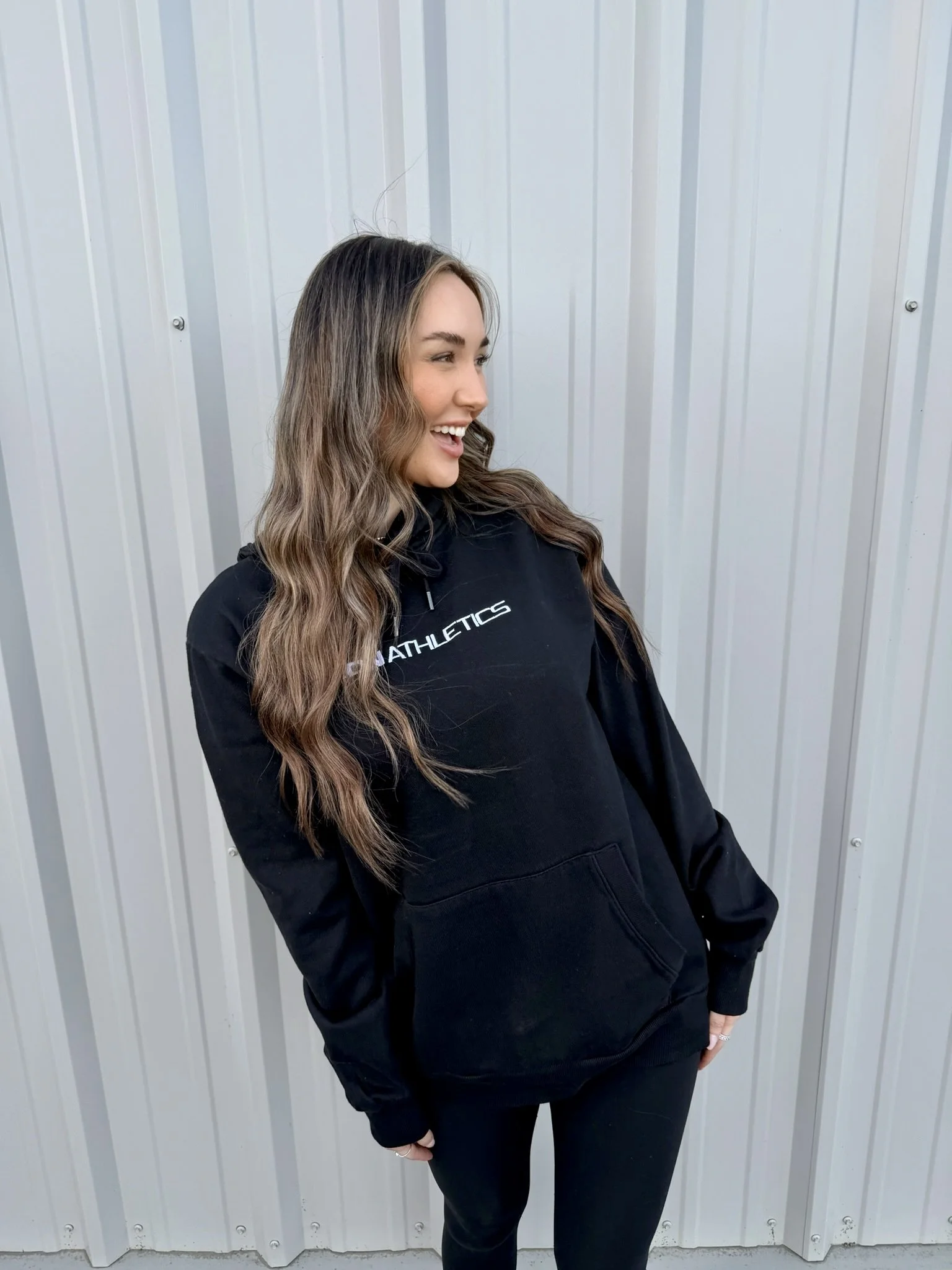 Young woman with long, wavy brown hair smiling, wearing a black hoodie with 'ATHLETICS' written on it, standing in front of a metal wall.
